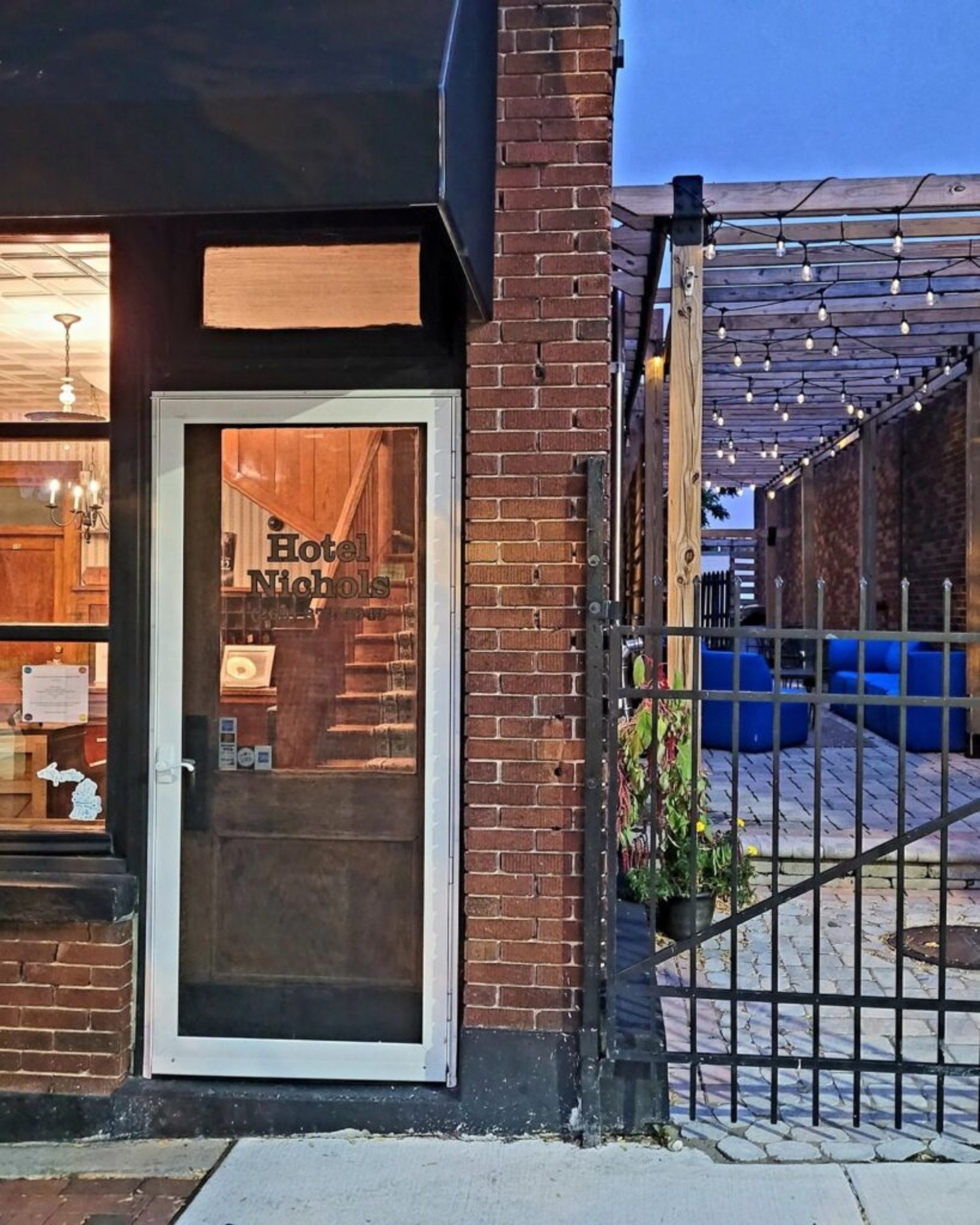 Entrance of Hotel Nichols with a glass door, brick facade, and adjacent patio area with string lights.