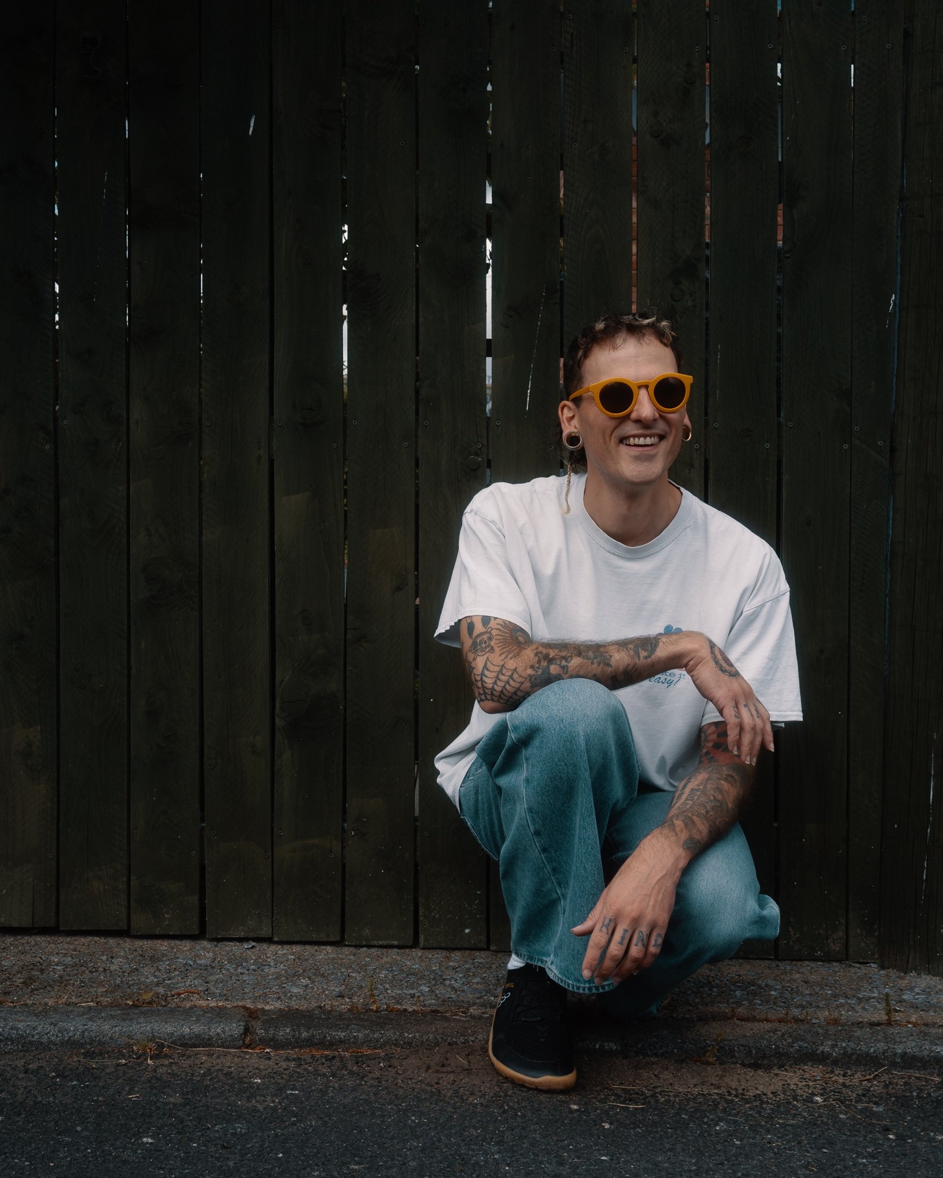 Cody wearing yellow sunglasses, a baggy white t shirt, and baggy jeans. They are crouched in front of a green wooden fence