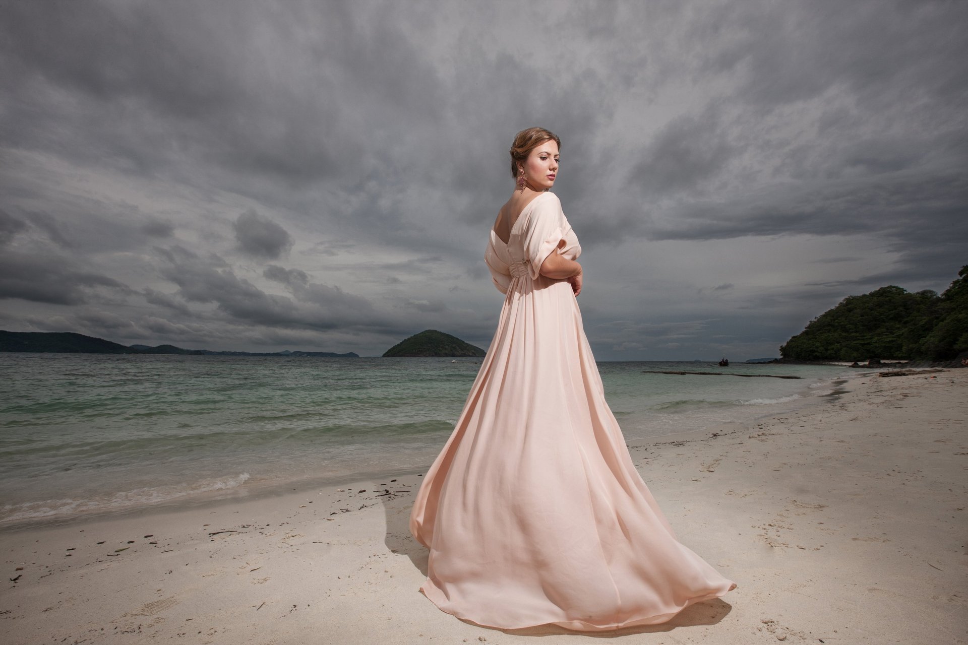 Elegant woman in flowing pastel dress on Madeira beach, Atlantic coast under dramatic cloudy sky.