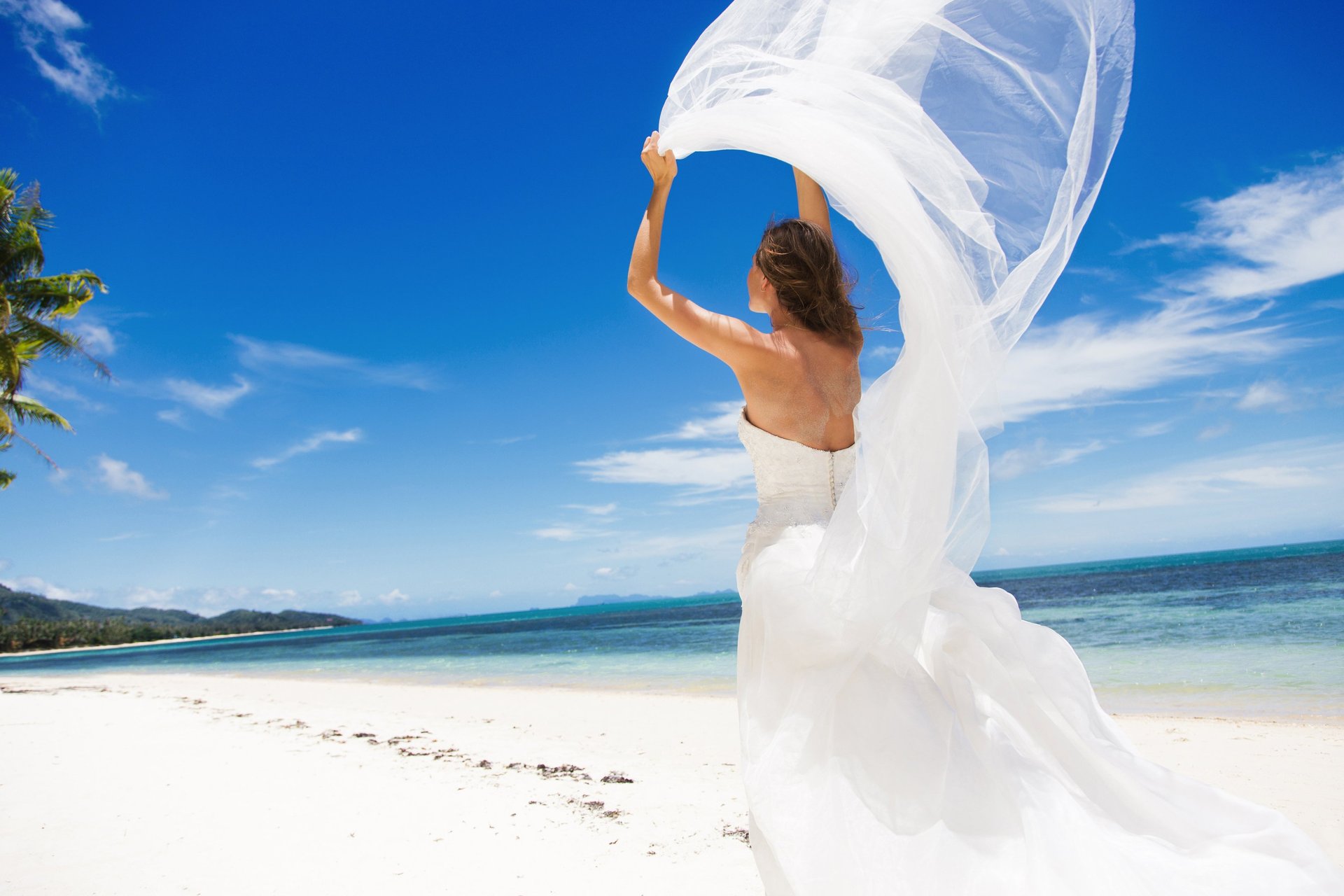 Beautiful bride in a white wedding dress holding veil on a sandy beach with turquoise ocean and blue sky in Madeira, Portugal
