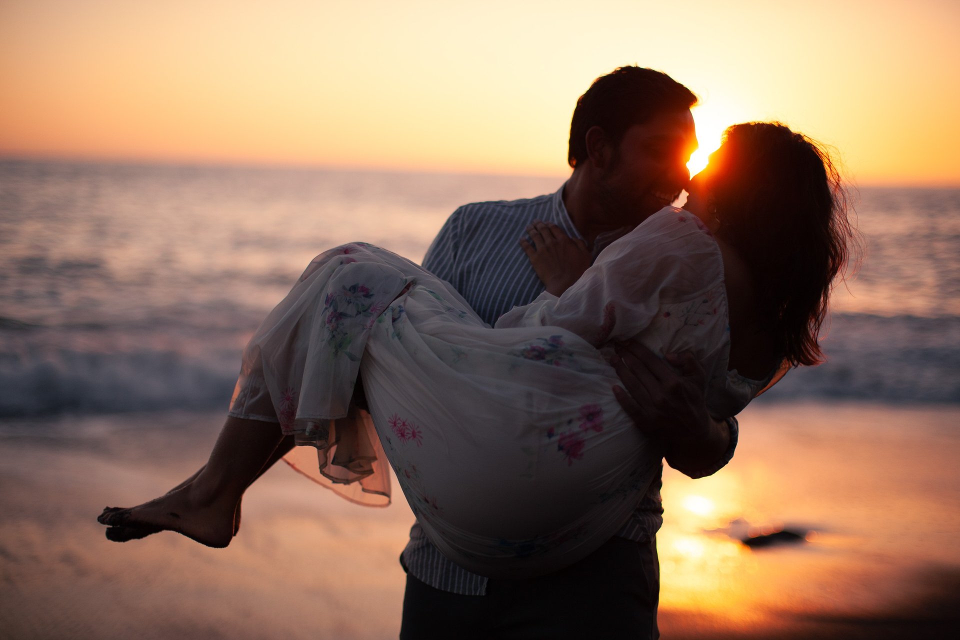 Man lifting kissing woman at sunset on Praia Formosa black sand beach, Madeira, ocean waves, romantic couple scene.