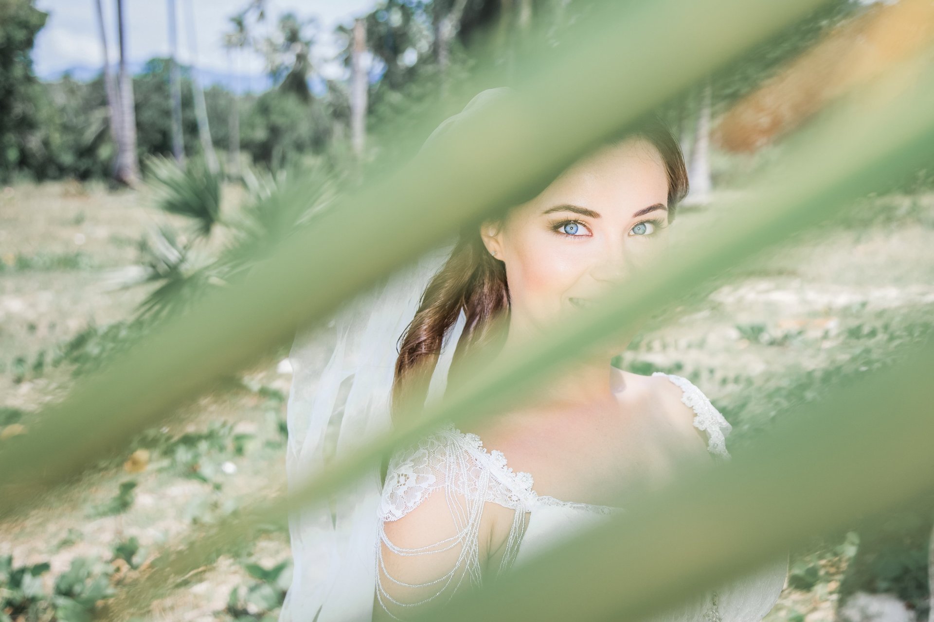 Bride with bright blue eyes looking at the camera through soft blurred palm leaves in the Island landscape of Madeira.