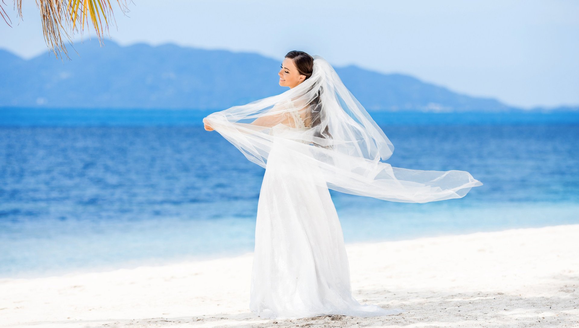 Bride in white dress and veil on sandy Madeira beach with ocean and mountains.