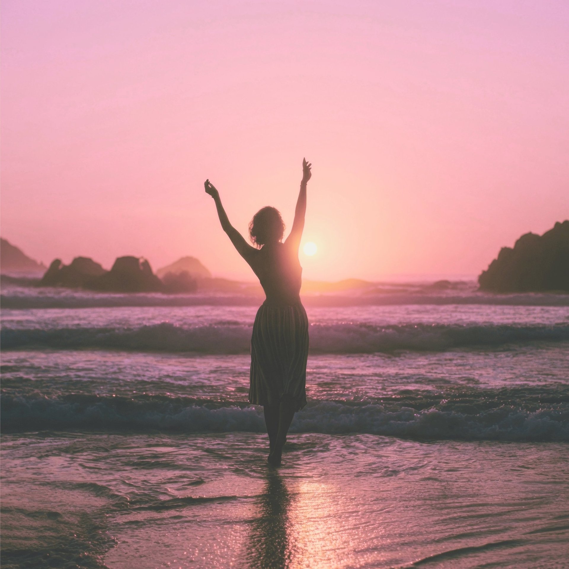 A woman standing on a beach embracing the pink sunset with her arms held up, feet in the ocean