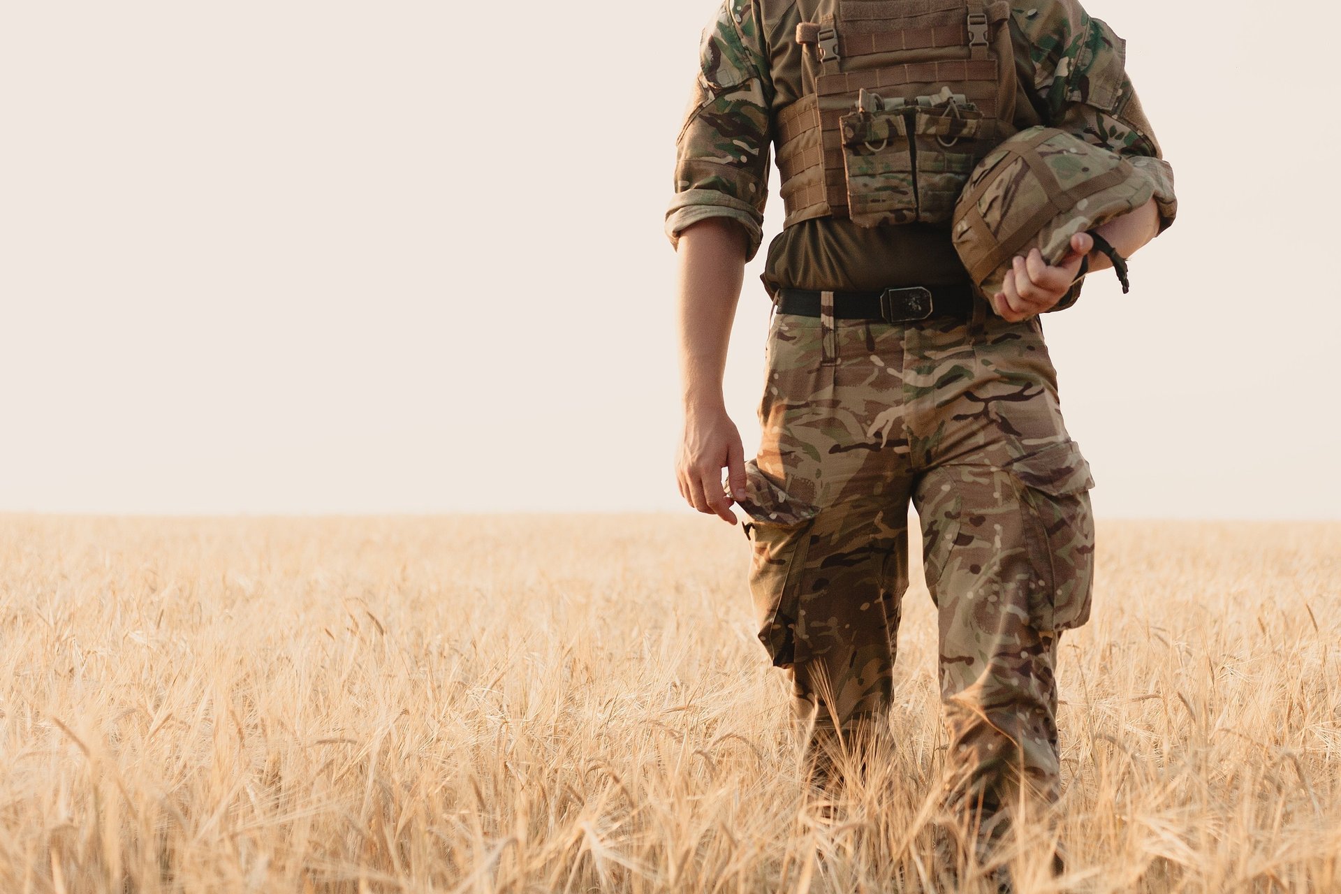 Soldier in camouflage walking through a golden wheat field, holding a helmet. Serene, natural landscape.
