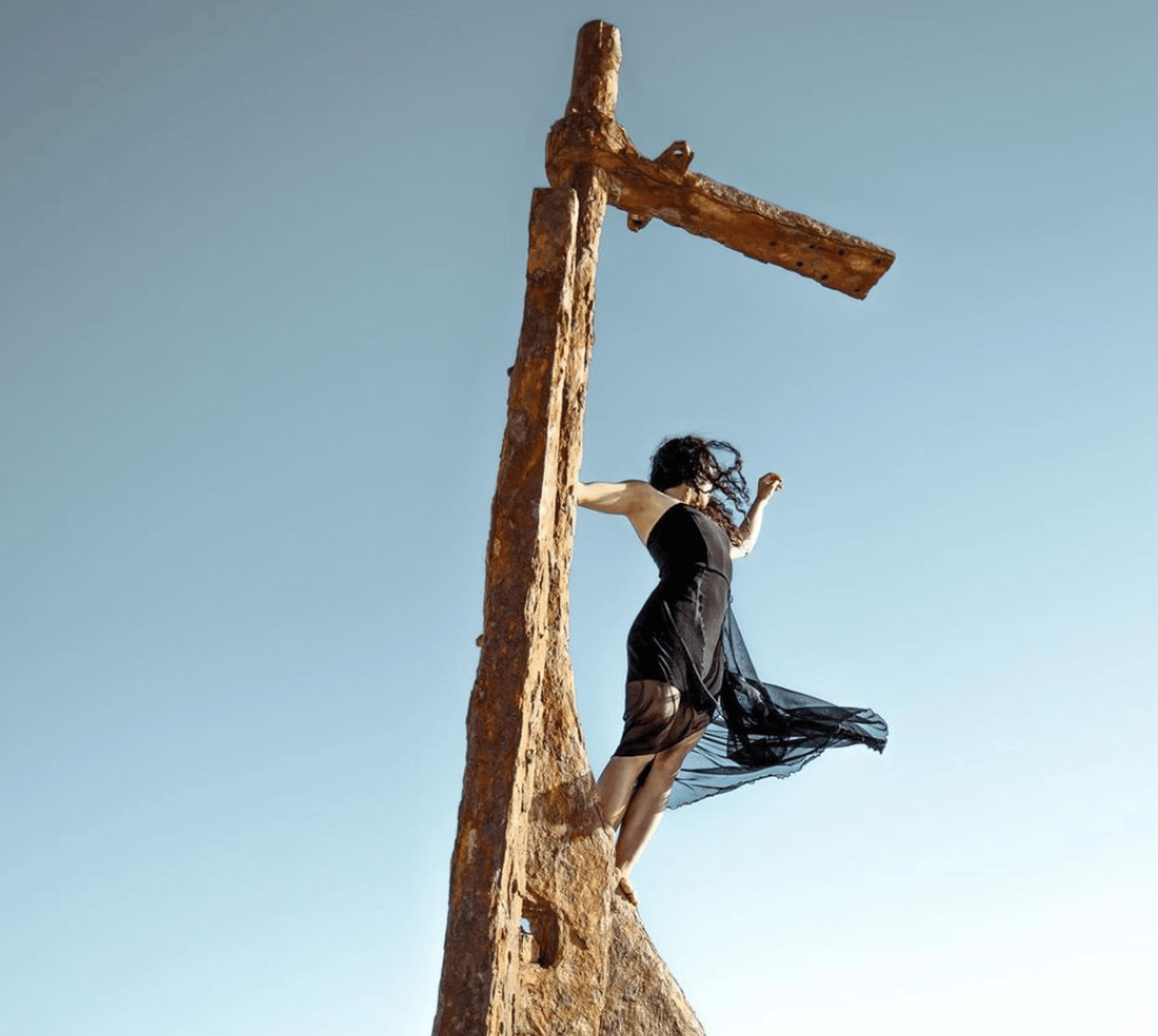 woman posing and stretching on the mast of an old rusted ship. She's wearing a black dress which is blowing in the wind. 