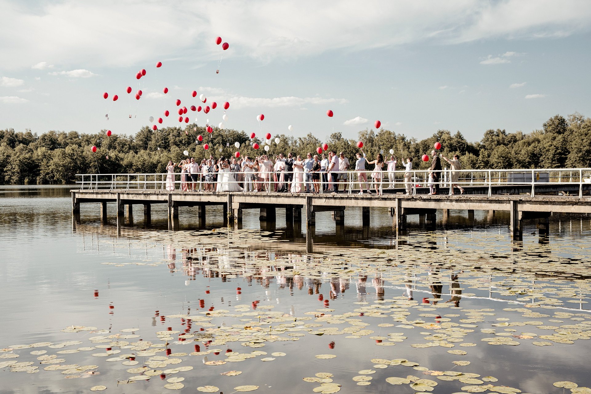 Hochzeitsgesellschaft lässt rote Luftballons auf einem Steg am See steigen – Gruppenmoment einer Hochzeitsreportage.