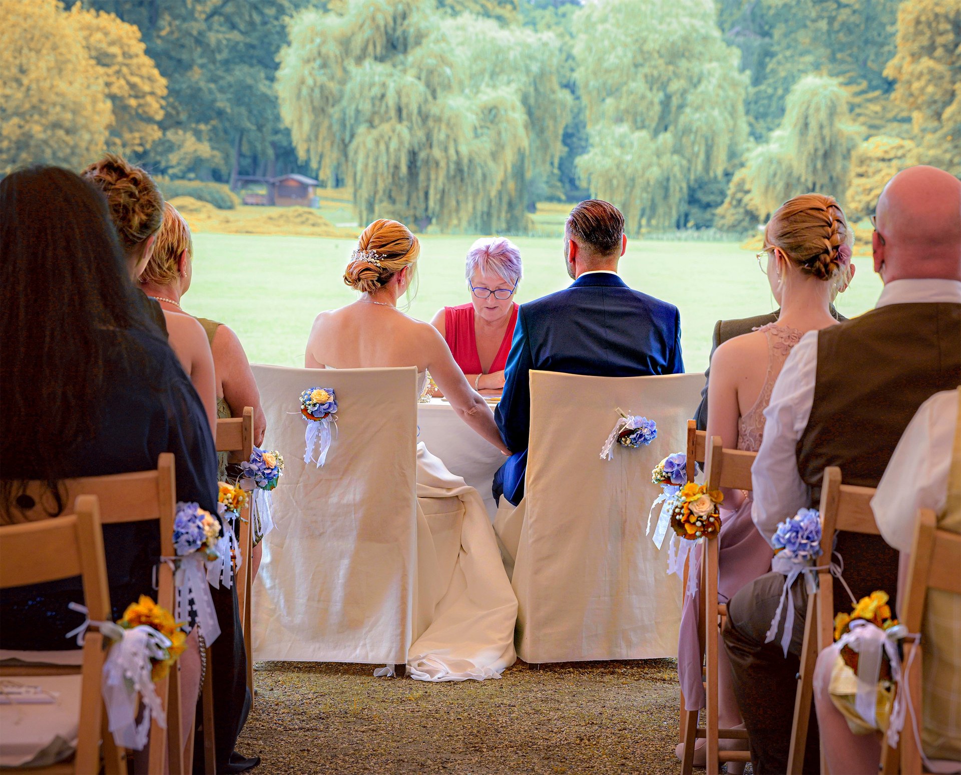 Brautpaar sitzt während der Trauung an einem Tisch, umgeben von Gästen bei einer freien Trauung im Grünen.