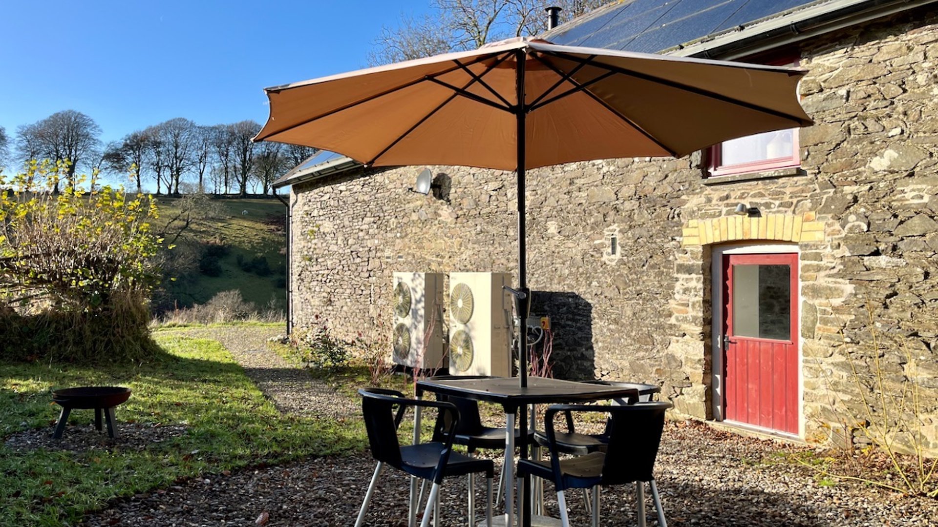 a table and chairs with parasol in a garden with stone building behind