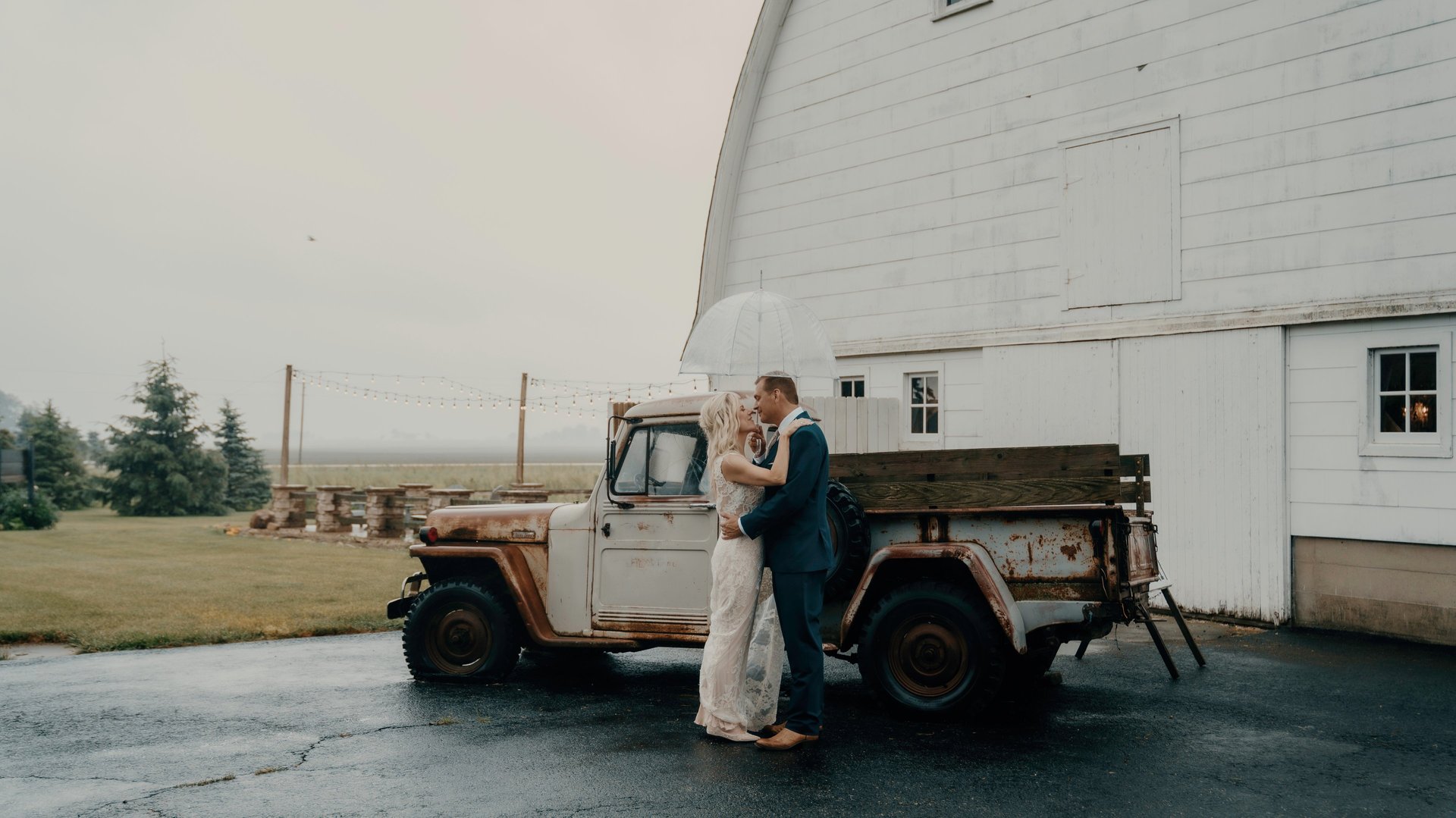 Bride and groom kissing under an umbrella by a vintage truck at rustic barn wedding – captured by Anngye & Juan wedding photo