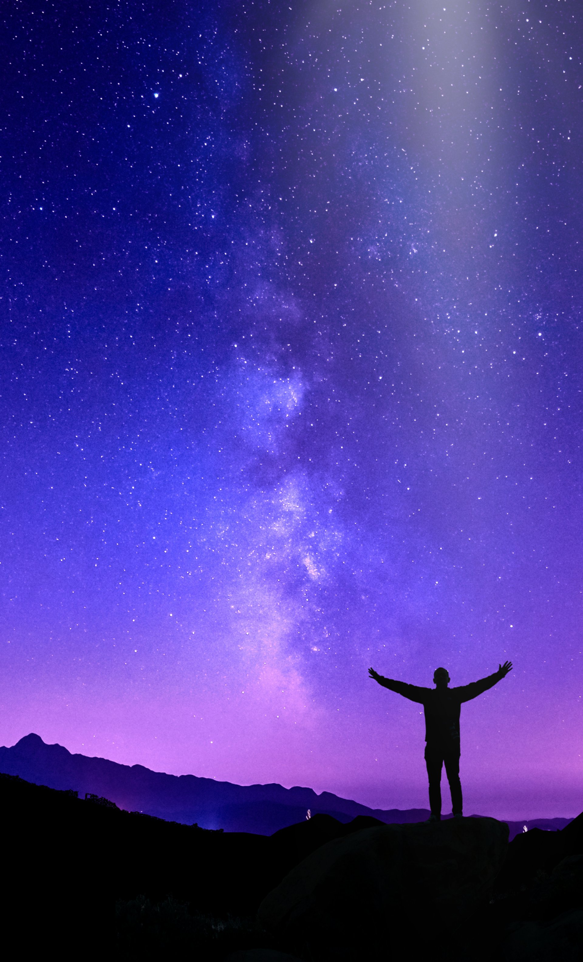 silhouette of person on top of mountain pointing flashlight on sky filled with stars at night time