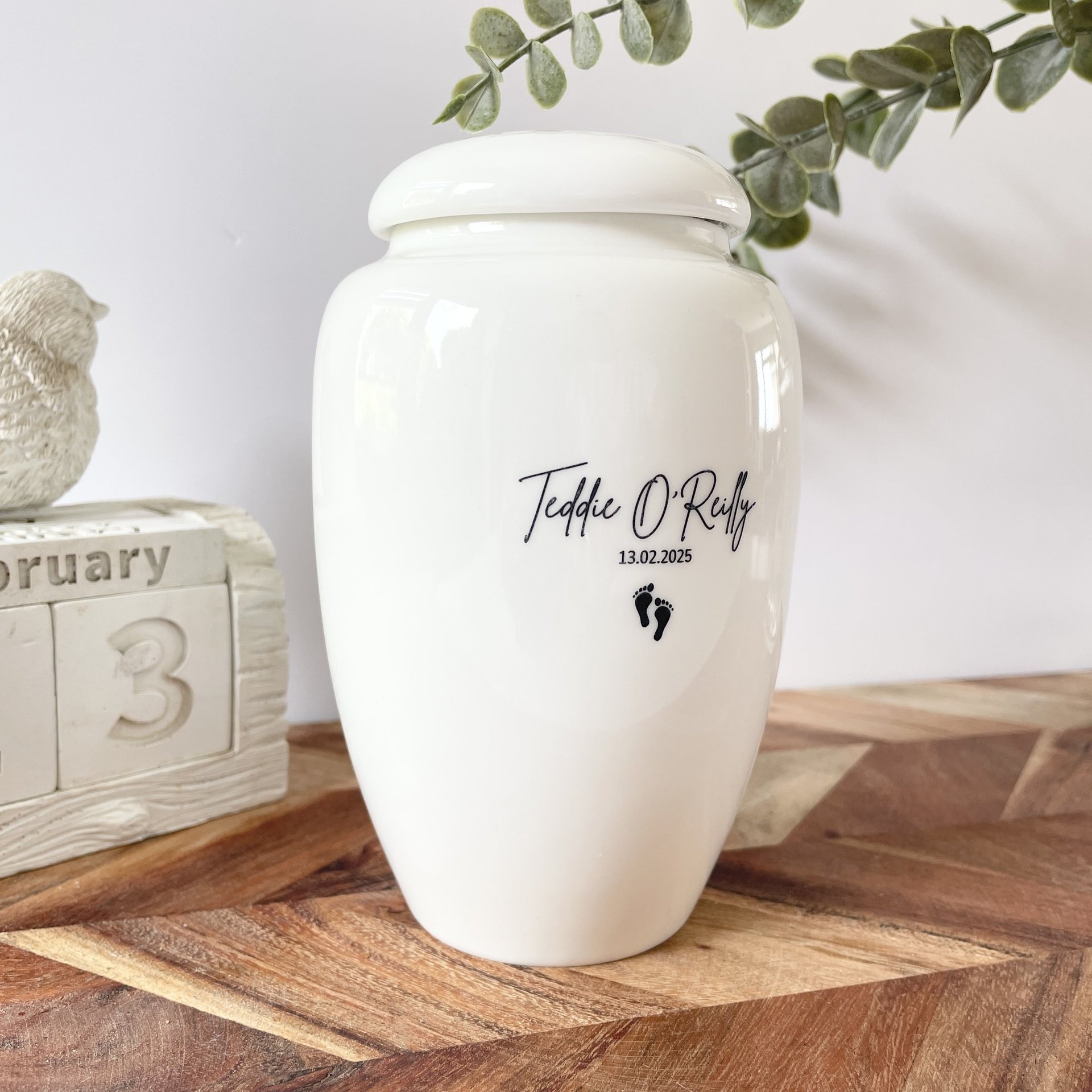 Elegant white ceramic urn with glossy finish, black script text "Teddy O'Reilly” and a small footprints icon