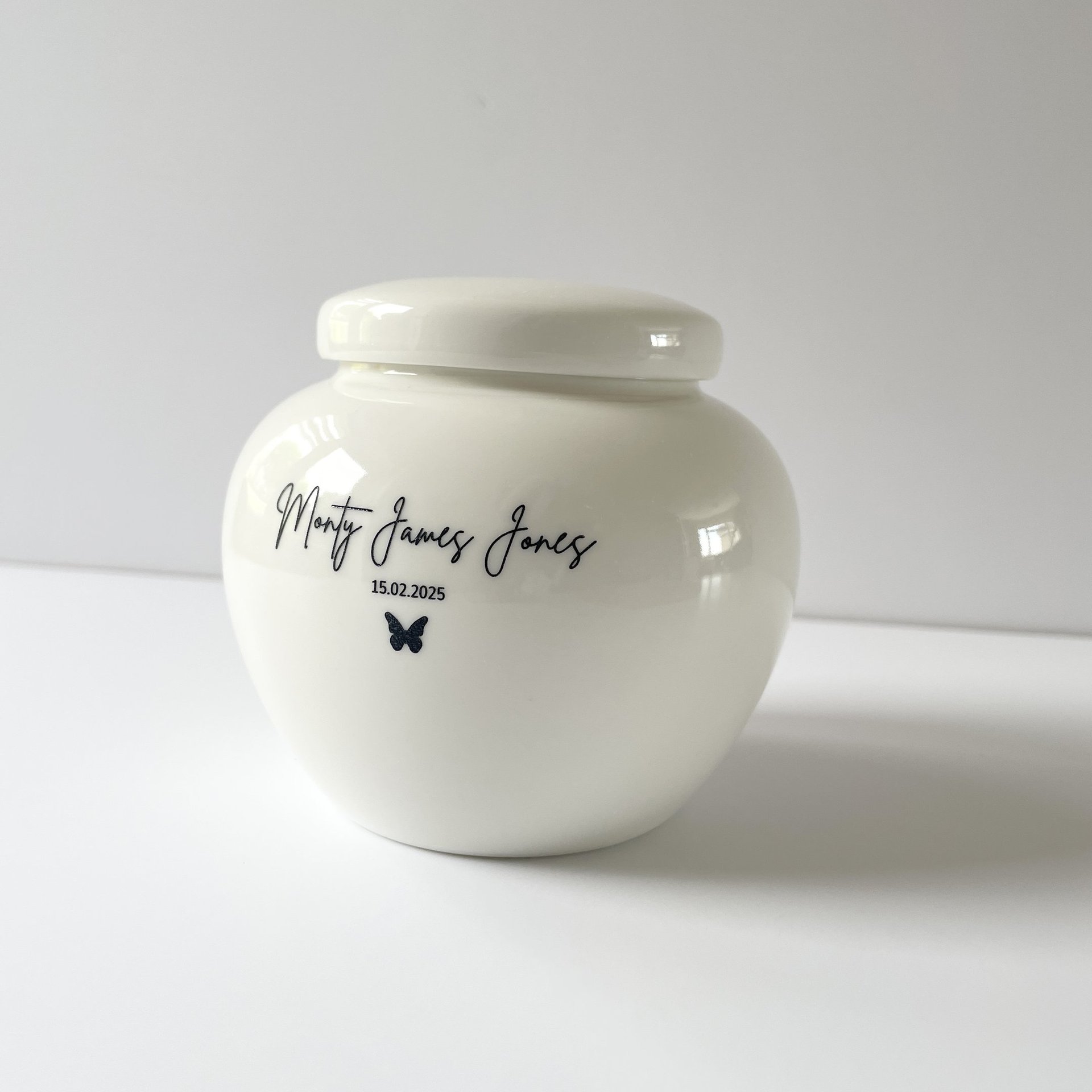 Rounded white ceramic urn with glossy finish, black script text “Monty James Jones” and a small butterfly icon.