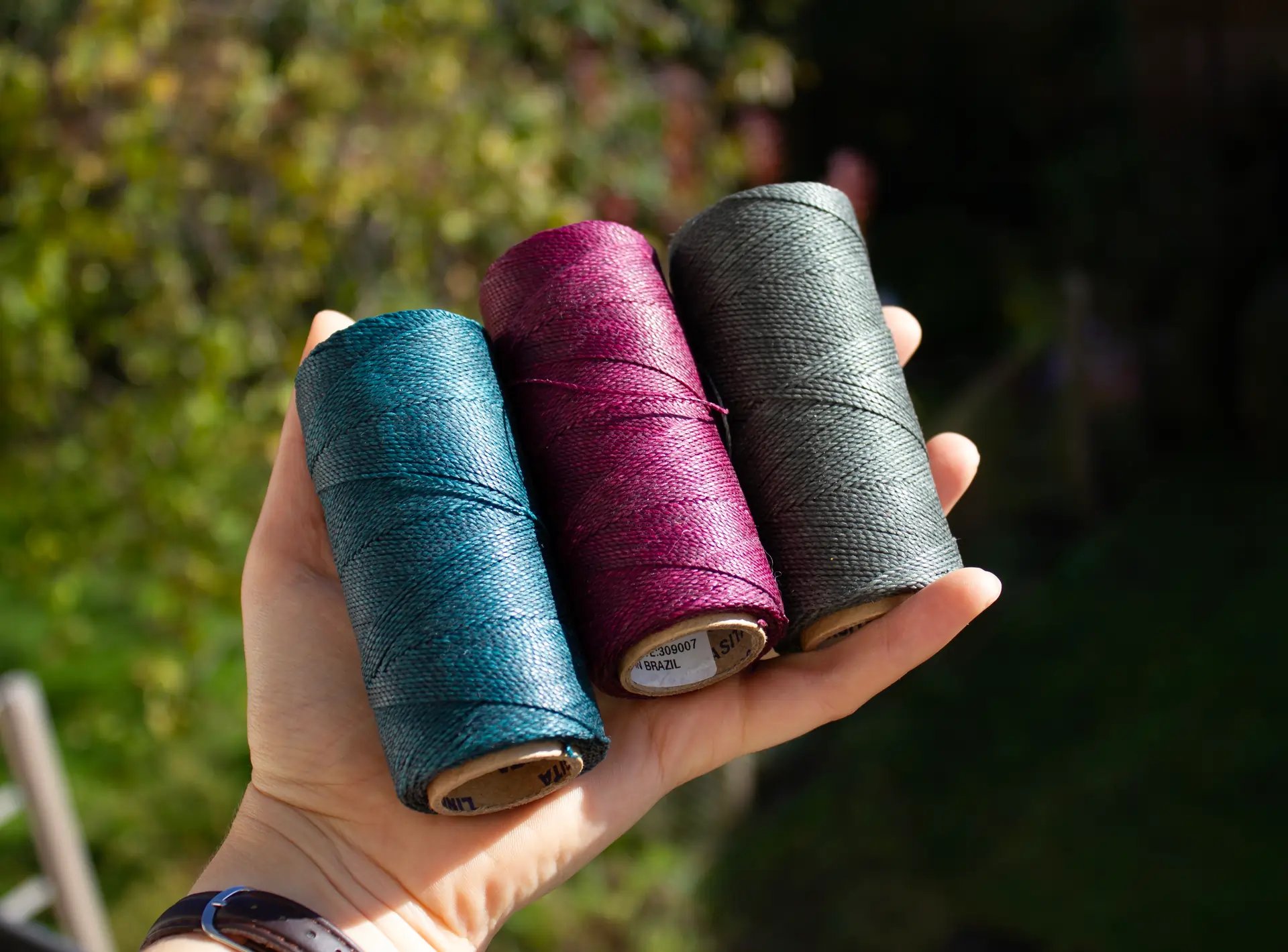 Hand hält drei Garnrollen in Türkis, Lila/dunkelrot und Dunkelgrün/olive/olivgrün