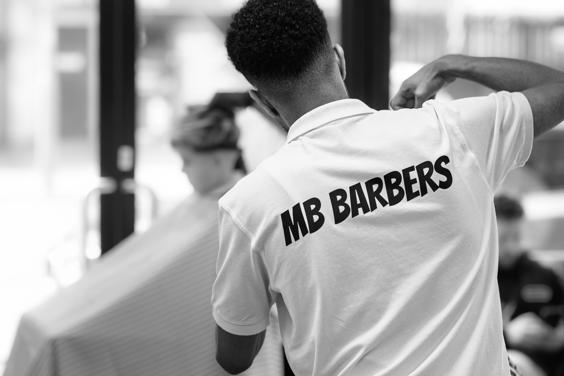 The back of a barber cutting a customers hair, with a blurred customer in the background