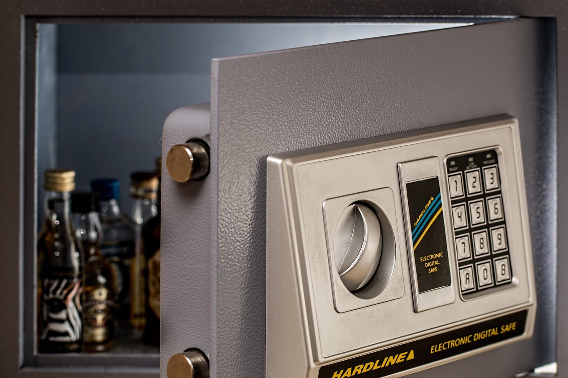 Open electronic hotel safe with keypad and dial, door ajar revealing miniature liquor bottles inside.