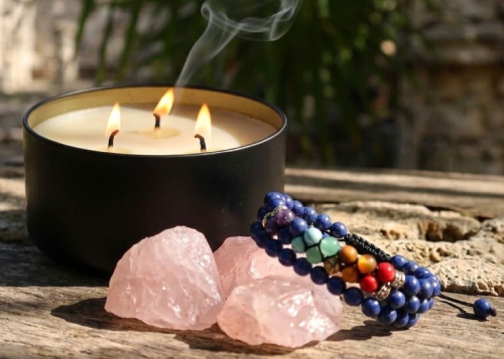 Vela encendida, cuarzos rosas y brazalete de chakras sobre madera rústica con fondo tropical de la Riviera Maya.