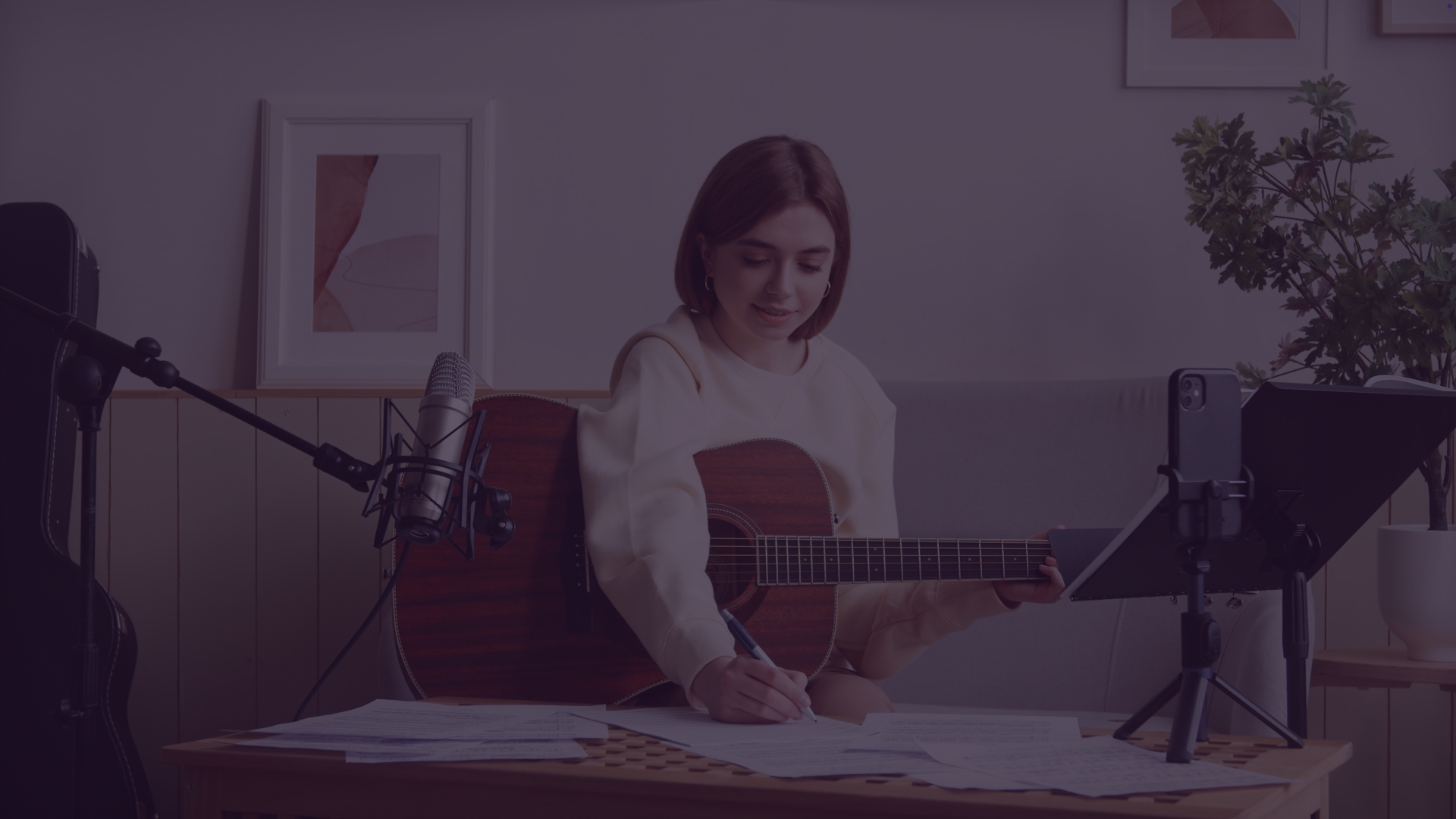 Young woman playing guitar on bed with laptop.