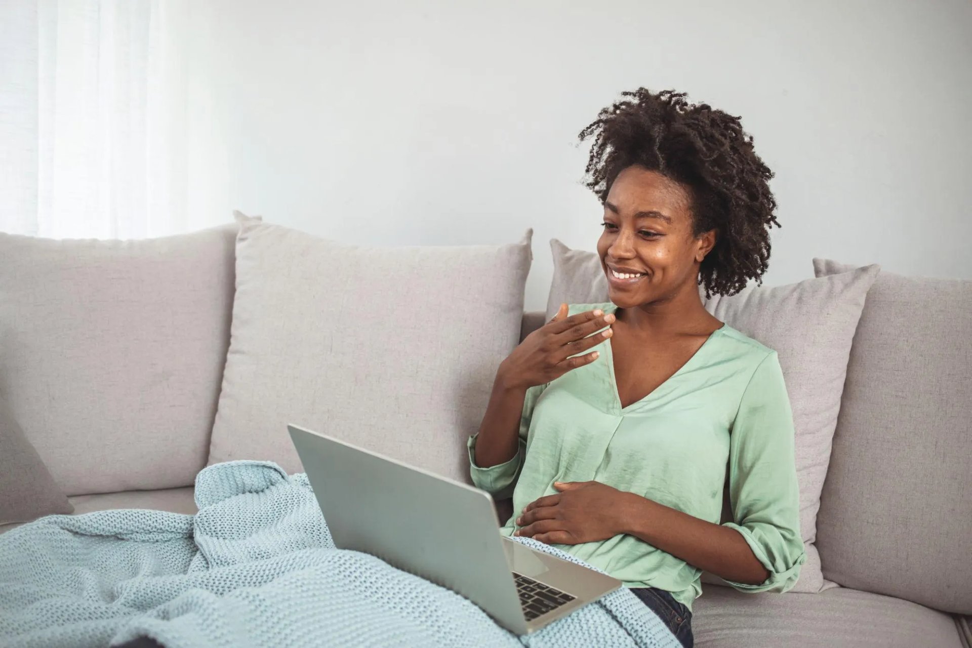 Femme souriante en visioconférence sur ordinateur portable, assise sur un canapé dans un cadre calme