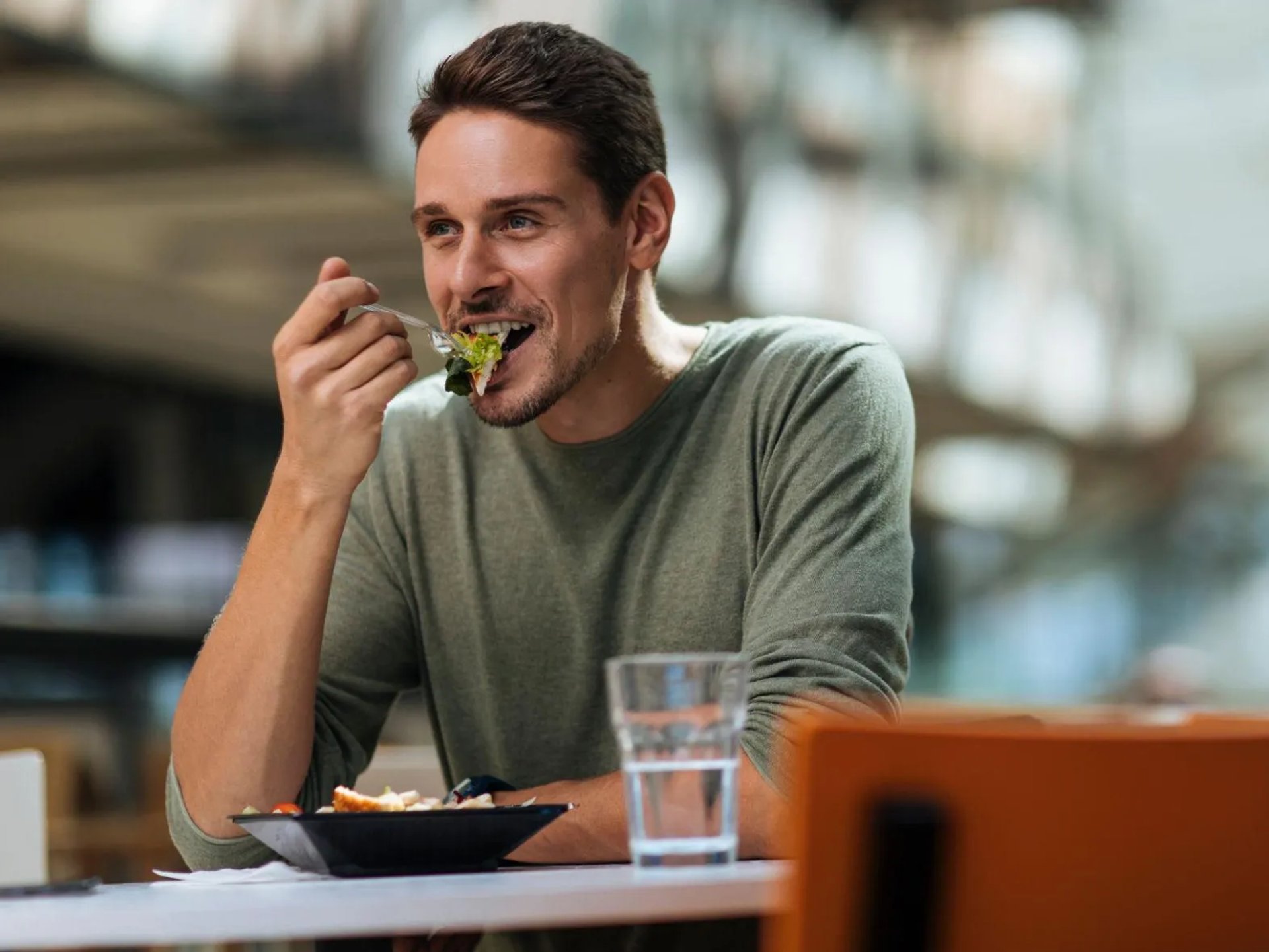 Homme mangeant une salade avec une fourchette, assis à une table, avec un verre d’eau devant lui.