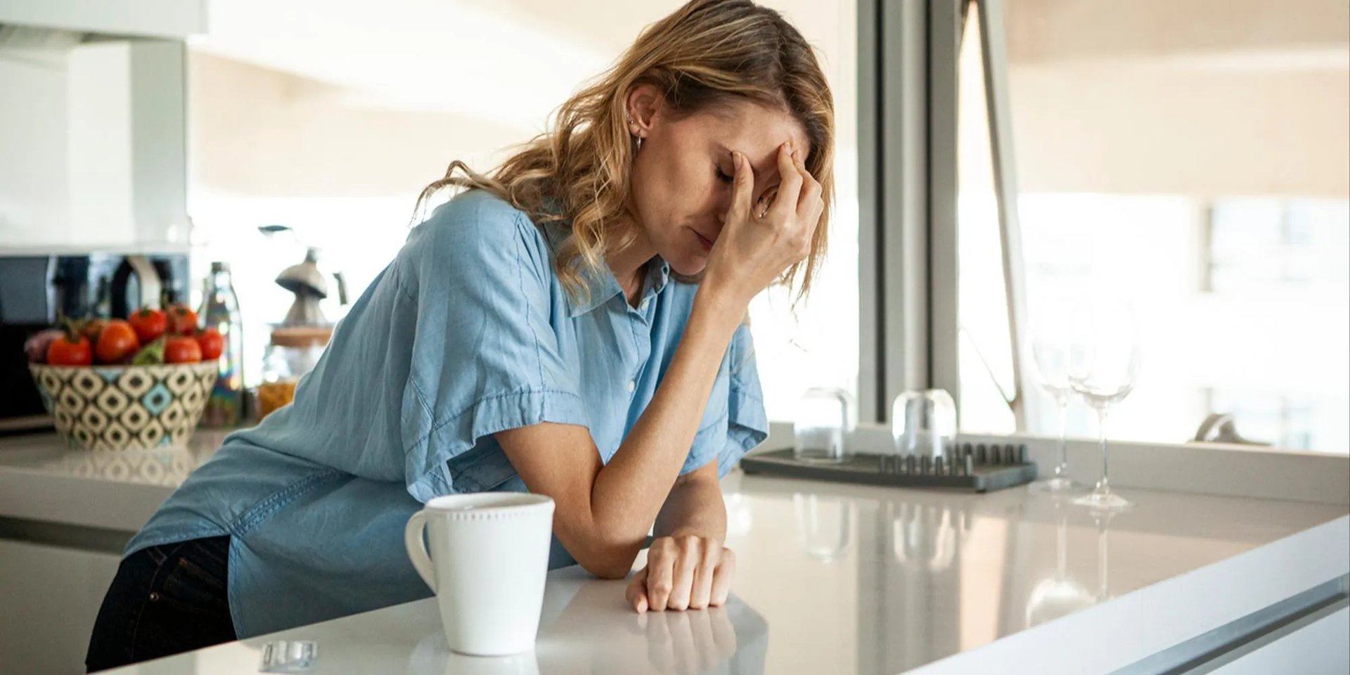 Femme fatiguée appuyée sur un plan de travail, main sur le visage, avec une tasse devant elle.