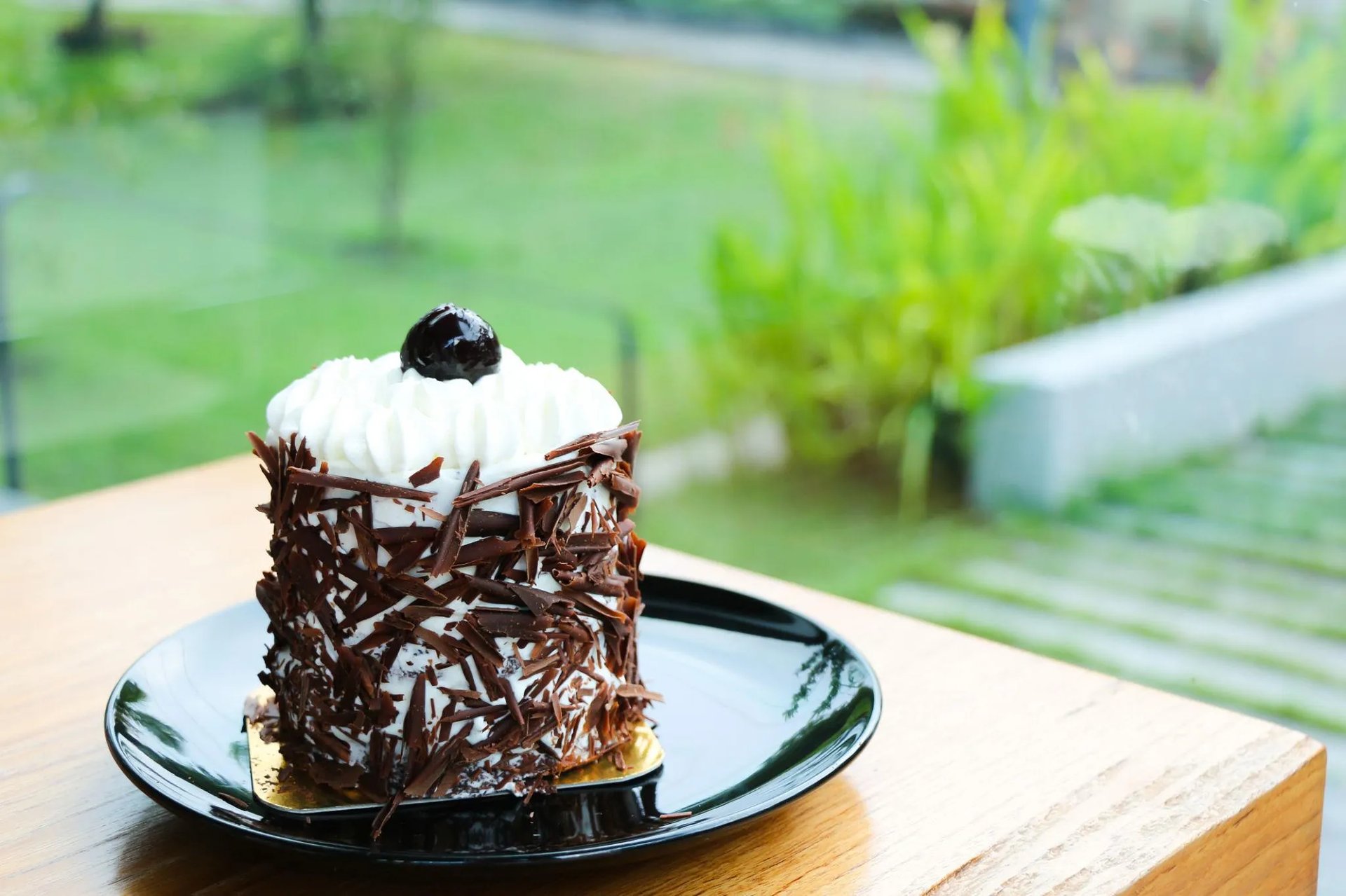 Part de forêt noire posée sur une assiette noire près d’une fenêtre avec vue sur un jardin.