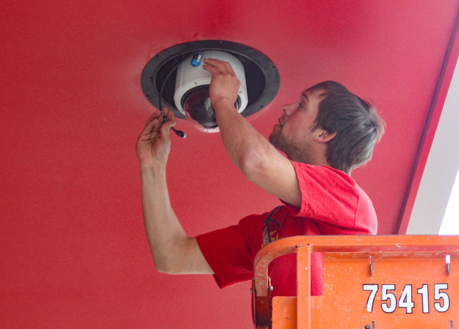 A technician on a lift installs a security camera into a red ceiling.