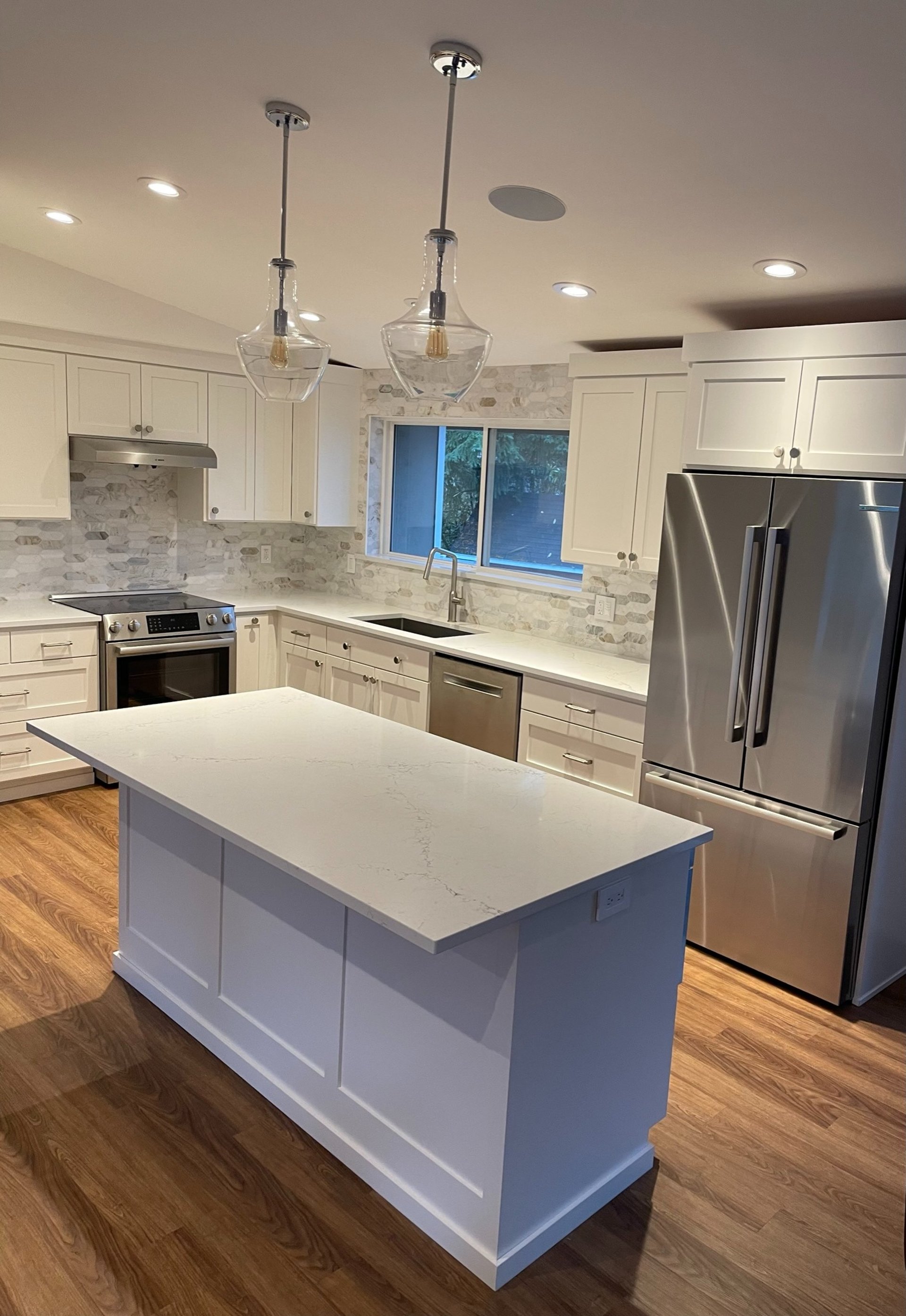 Completed kitchen remodel with white shaker cabinets, quartz island, stainless appliances, tile backsplash, and lighting.