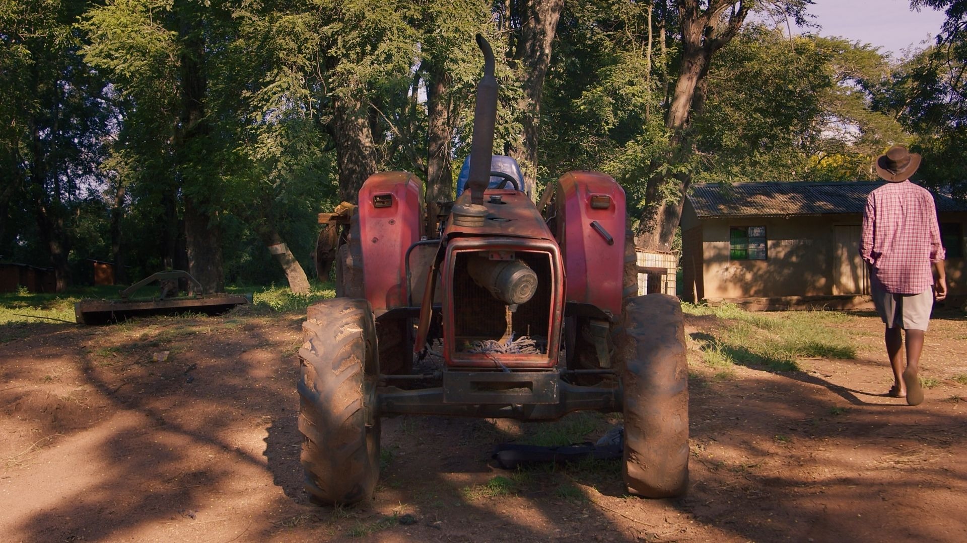 Farmer walking past a red tractor toward a farmhouse surrounded by trees on a rural farm.