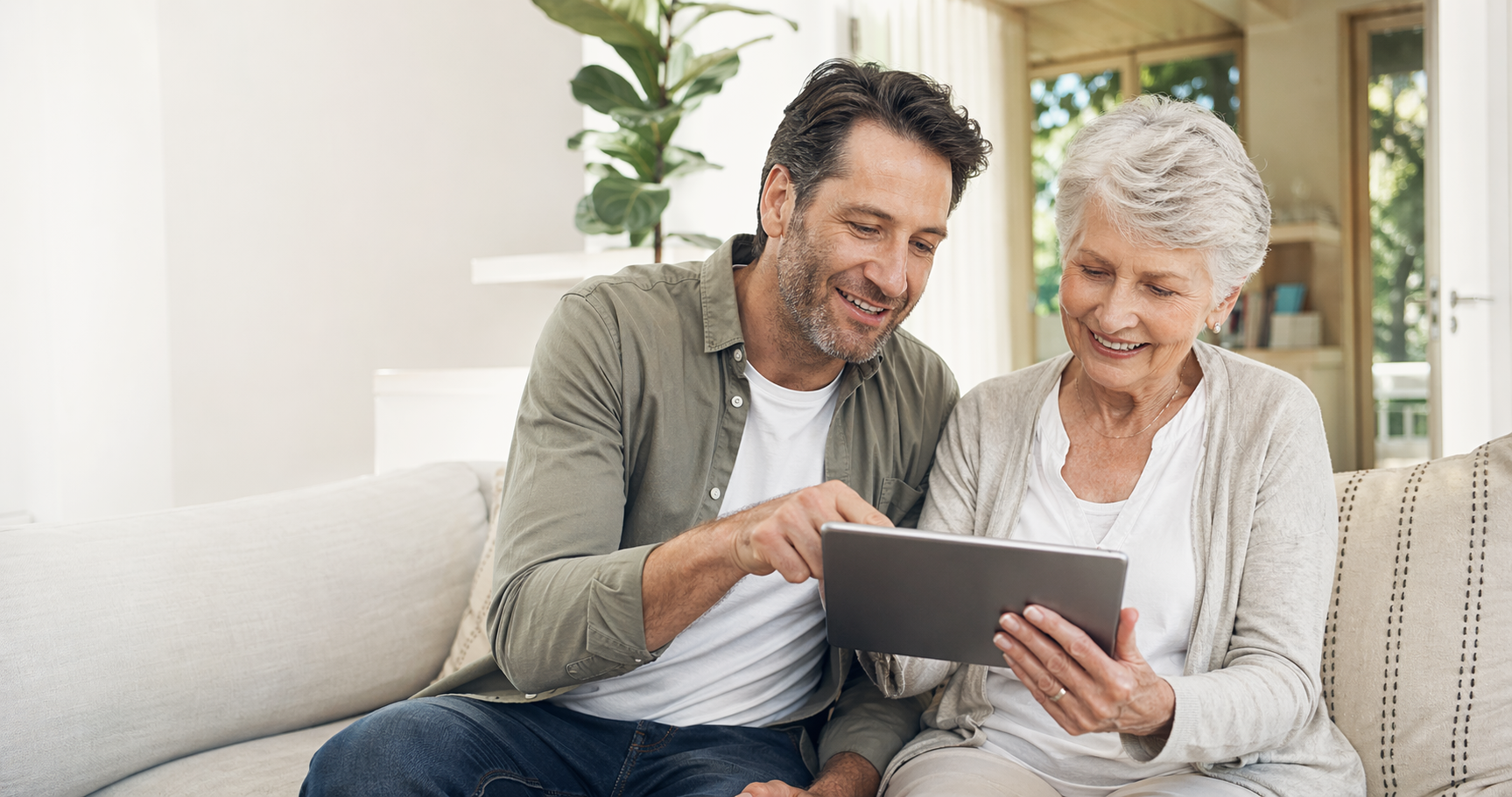 A smiling adult son and senior mother use a digital tablet together on a living room sofa.