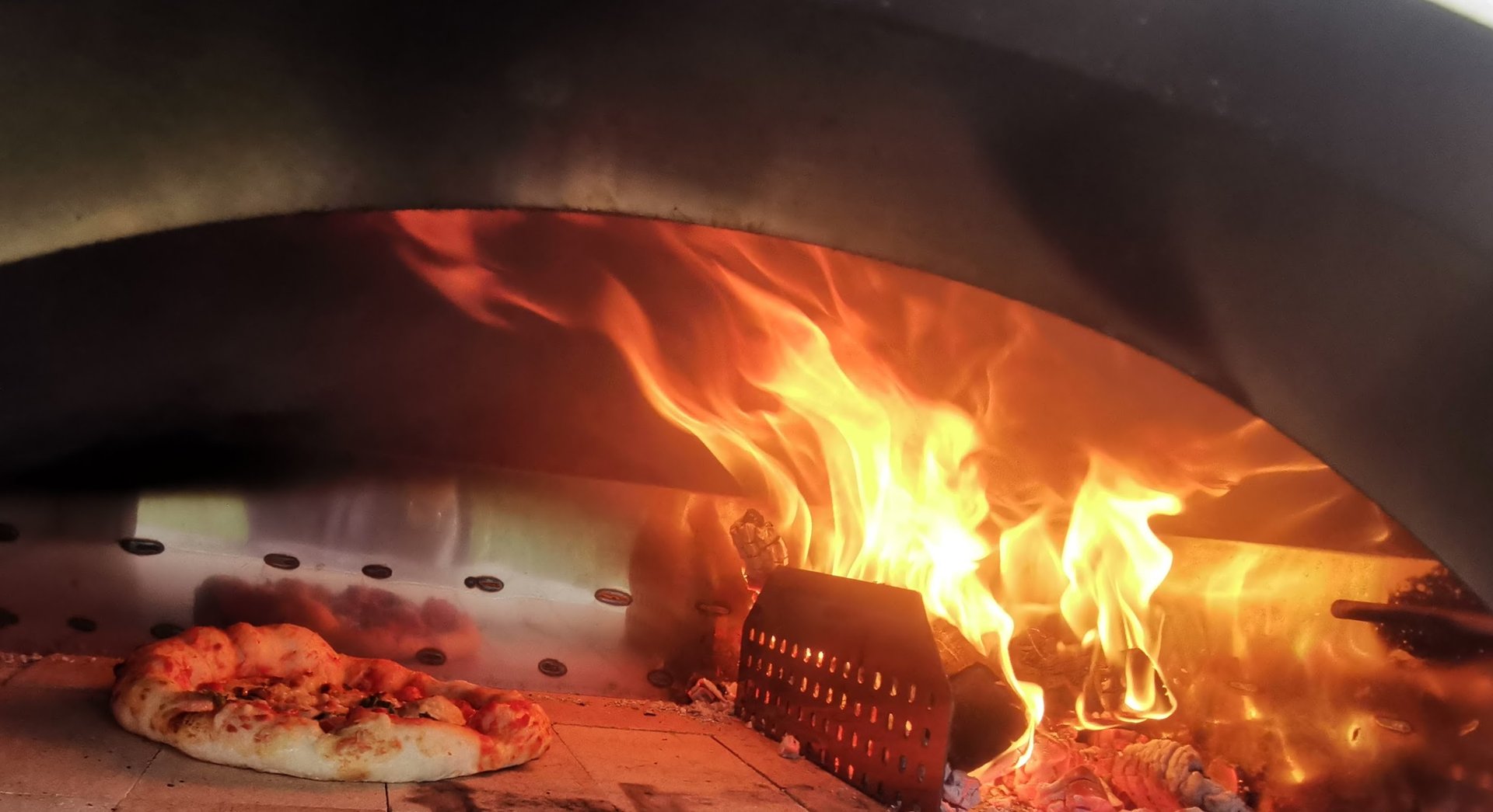 A fresh pizza baking inside a wood-fired outdoor oven with bright orange flames.