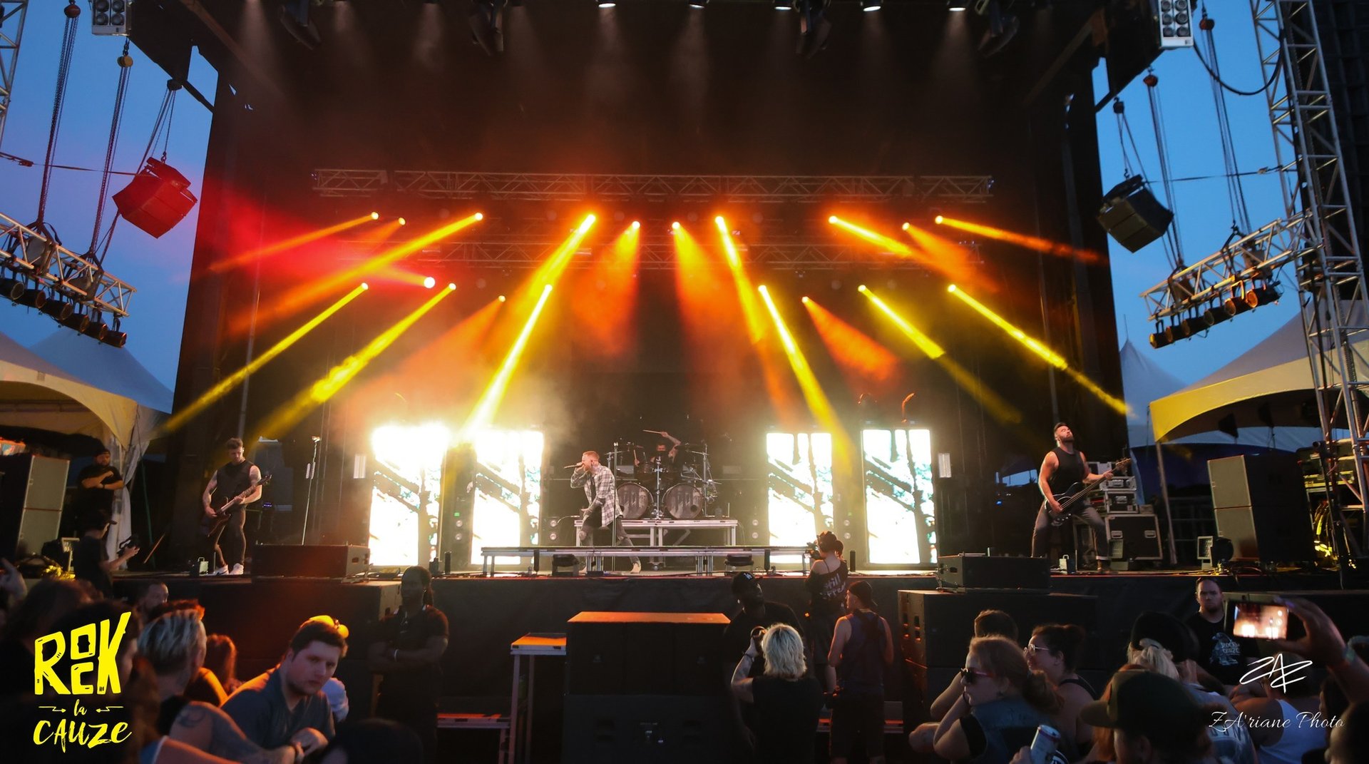 Live rock band performing on an outdoor stage with dramatic orange spotlights and a large crowd.