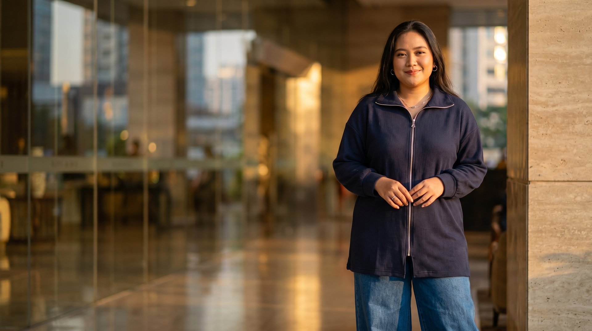 Smiling woman wearing a navy blue zip-up cardigan and jeans standing in a modern office building