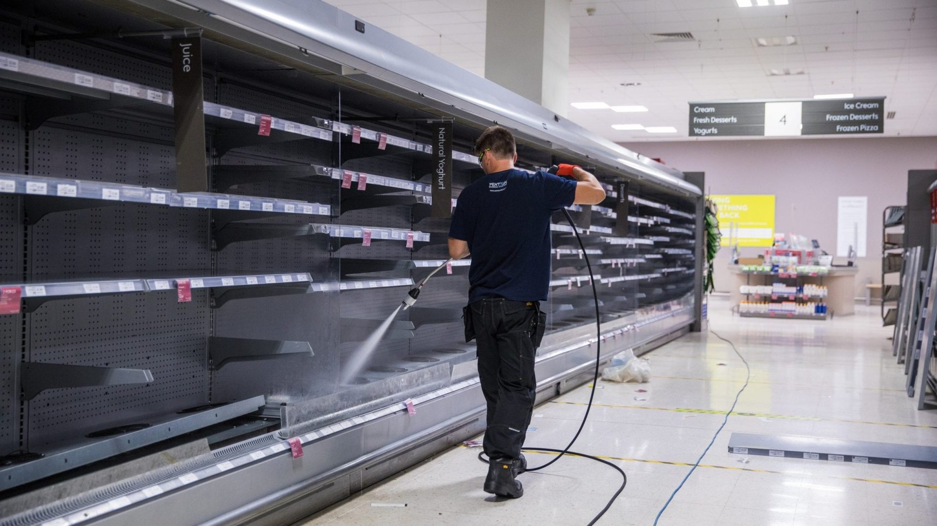A maintenance worker pressure washing empty refrigerated shelves in a supermarket aisle.