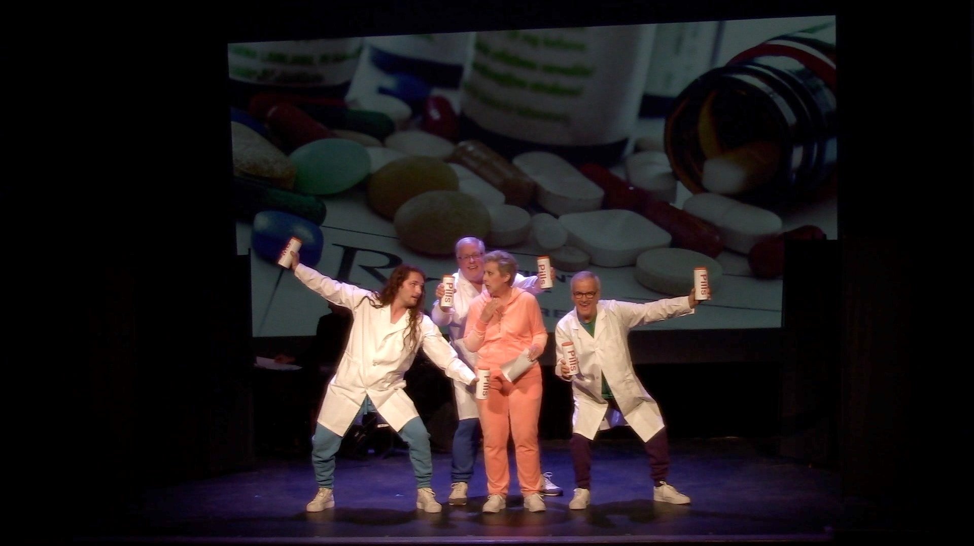 Performers in lab coats holding pill bottles dance in front of a pharmaceutical backdrop.
