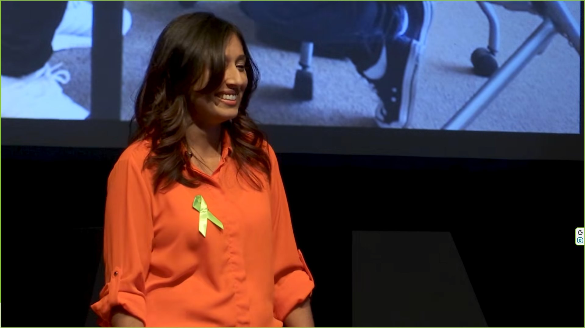 A woman in an orange shirt smiles on stage, wearing a green awareness ribbon, conveying positivity.