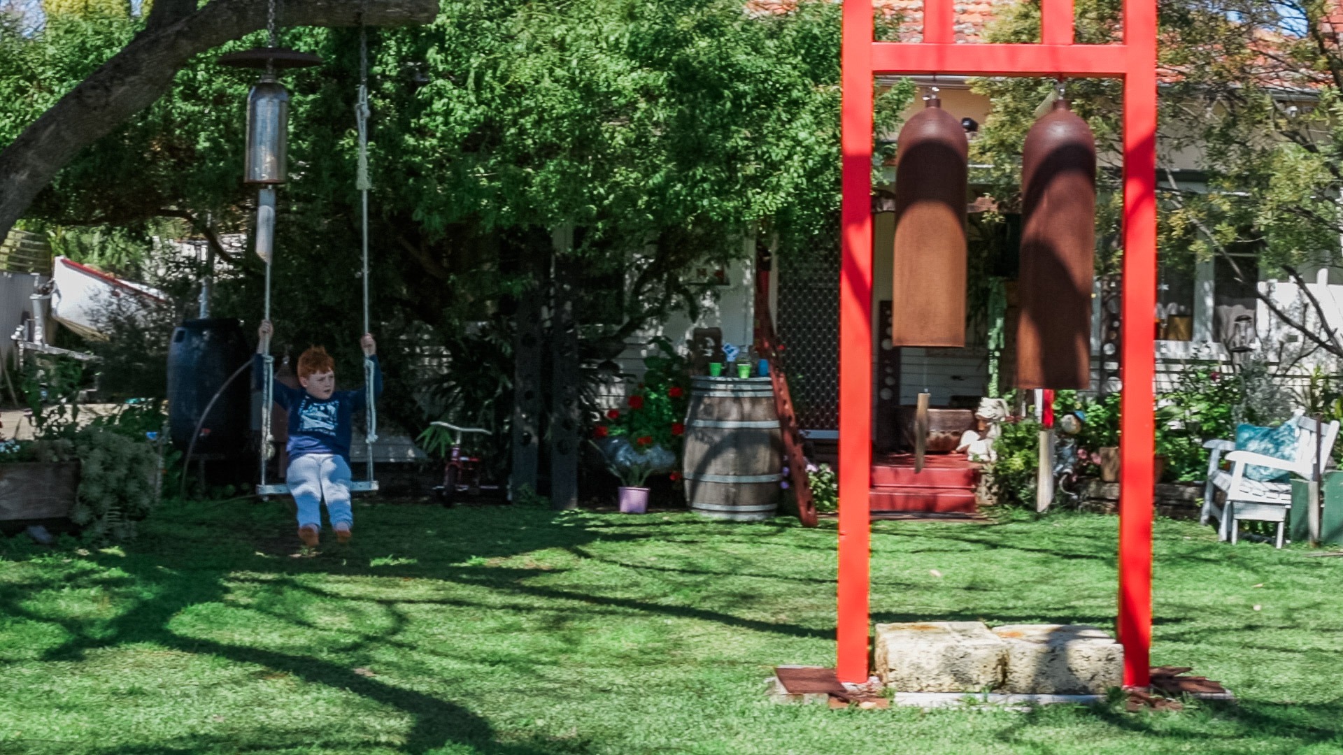 A young boy on a tree swing in a green garden with large hanging chimes and a red frame.
