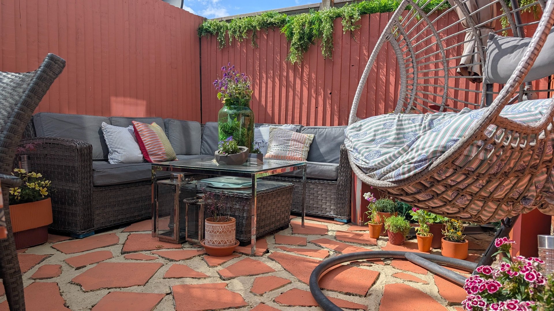 A cozy patio with a grey wicker corner sofa, hanging egg chair, and garden plants on terracotta tiles.