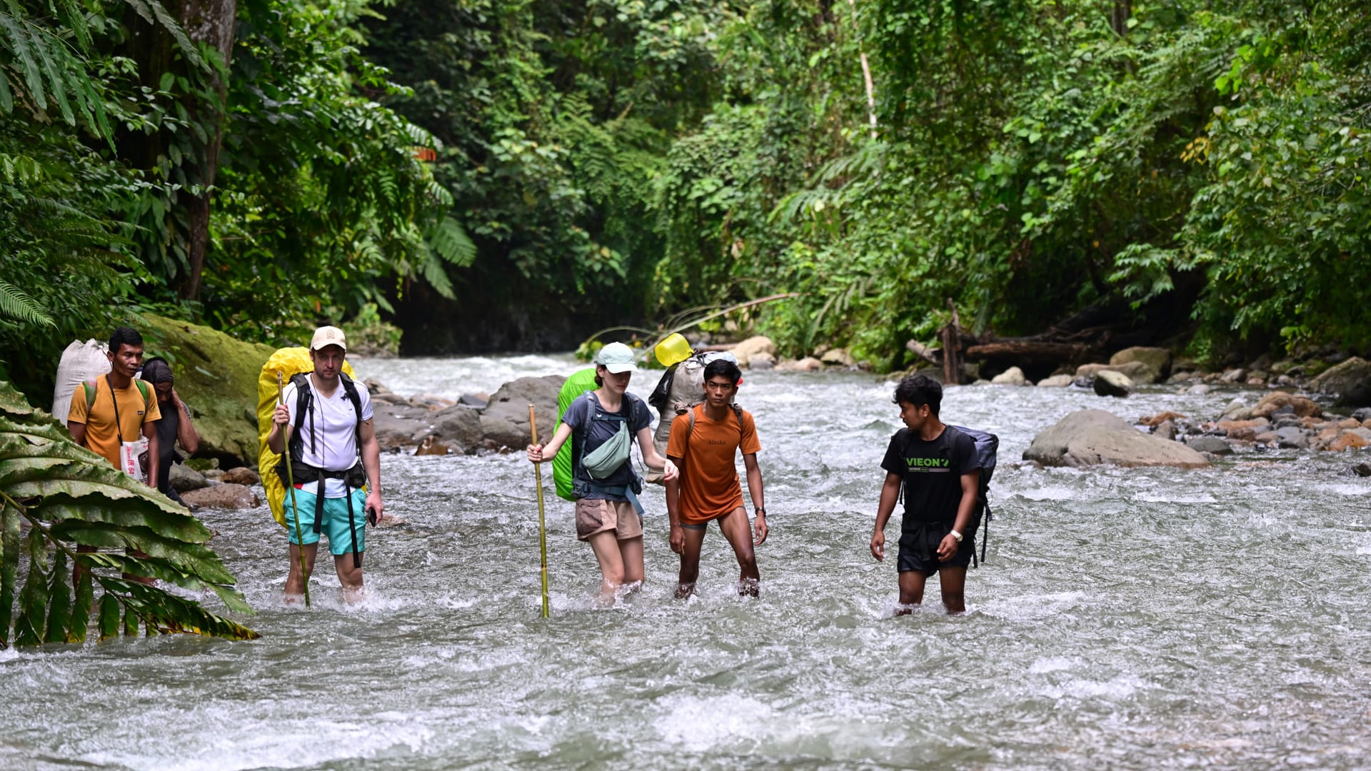 A group of hikers and local guides with backpacks cross a shallow river in a lush tropical rainforest.