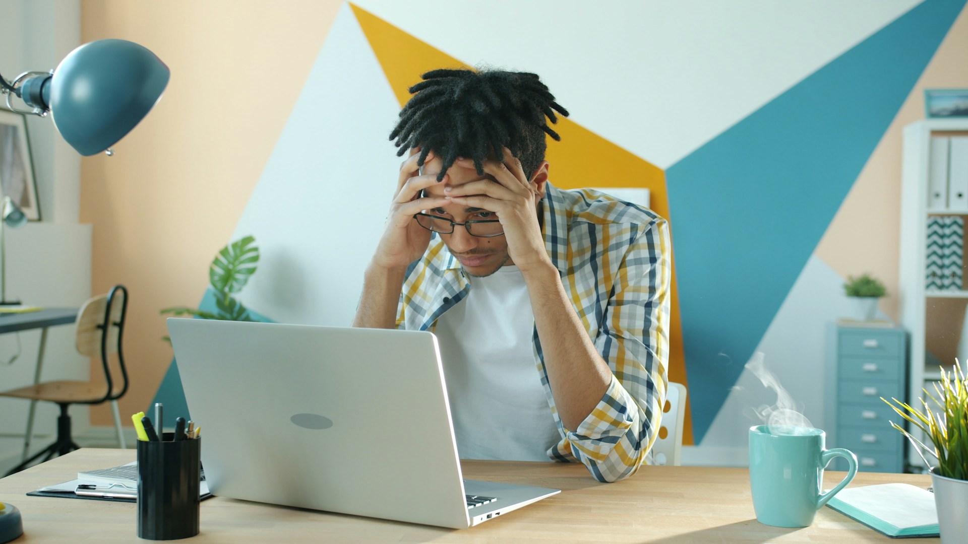 Frustrated young freelancer sitting at a desk with an open  laptop, holding his head in his hands.
