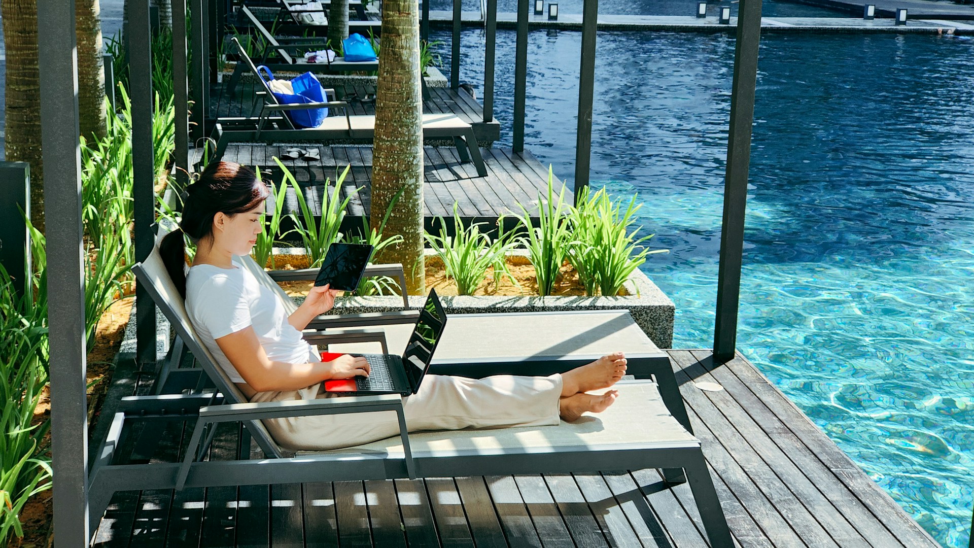 A digital nomad works on a laptop and tablet while lounging by a luxury resort swimming pool.