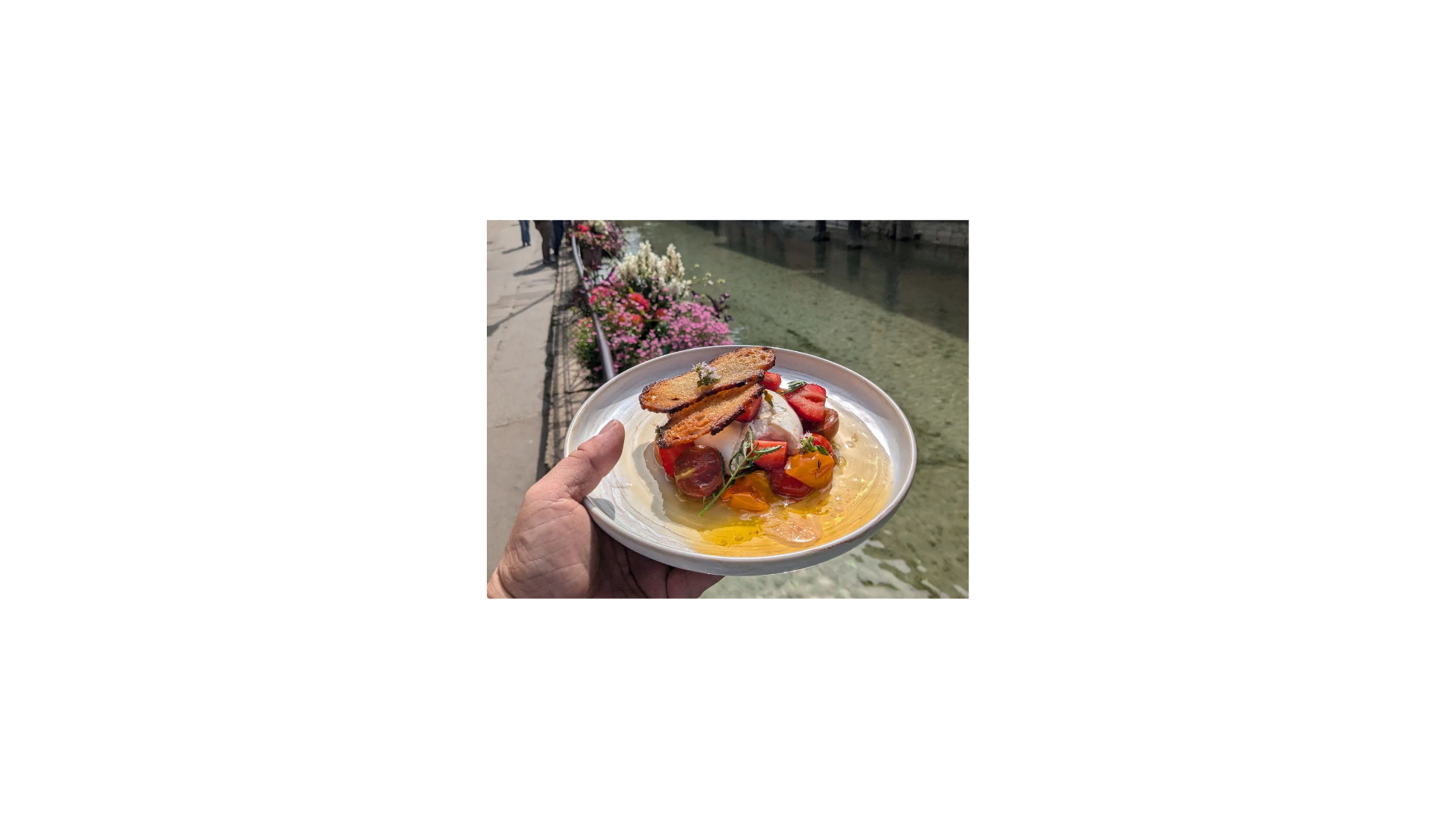 Une personne tient une assiette avec une burratta accompagné de fraises, tomates et un croustillant
