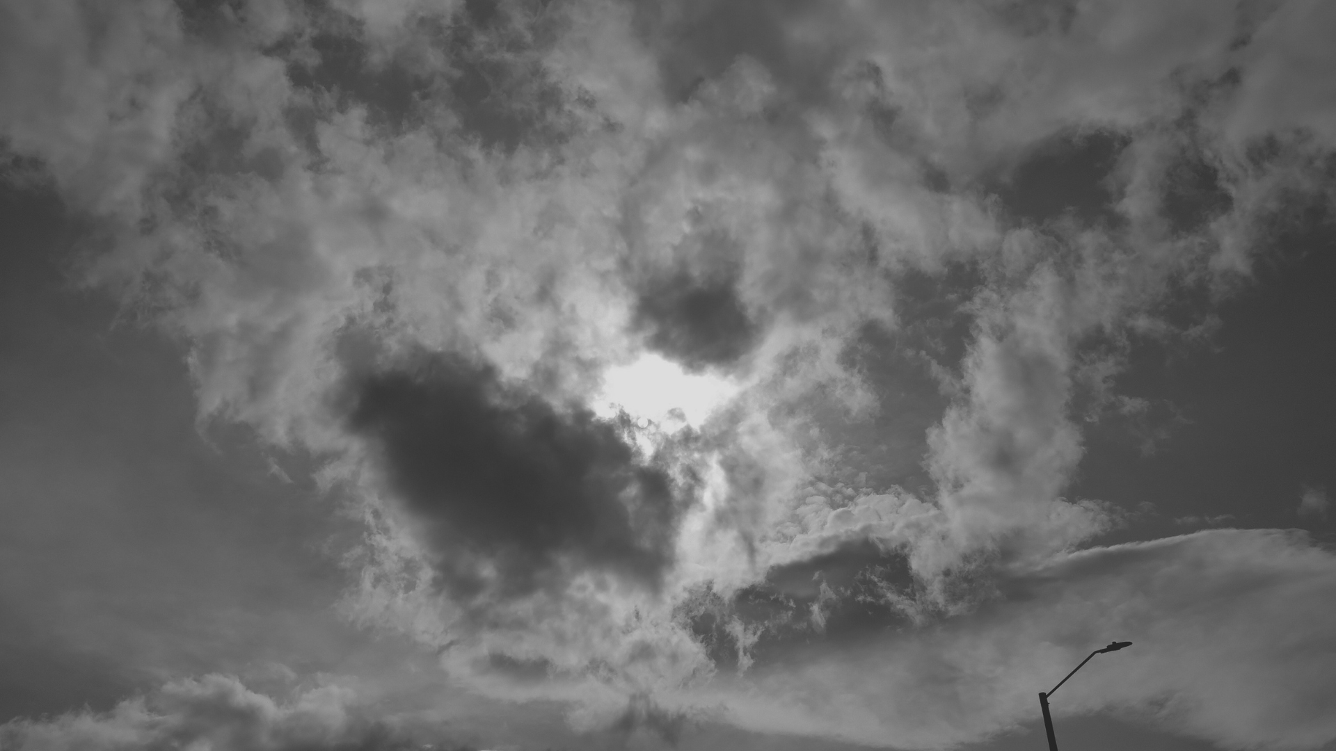 Cielo nublado en blanco y negro con luz entre nubes y poste urbano