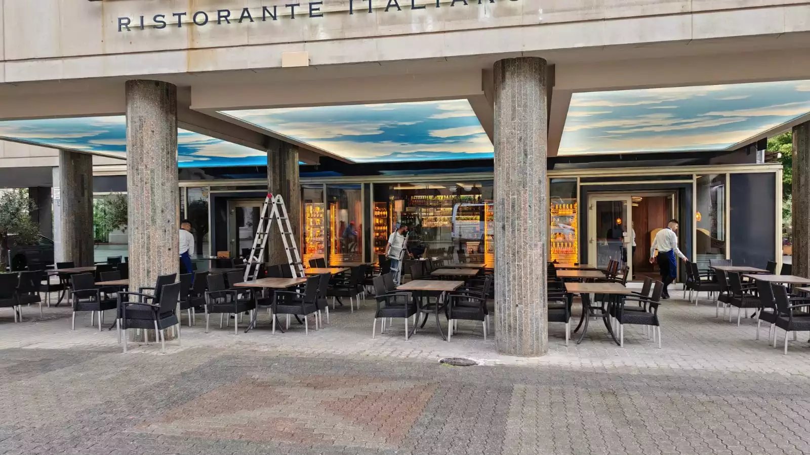 Outdoor patio of an Italian restaurant with modern chairs, stone pillars, and a cloud-painted ceiling.