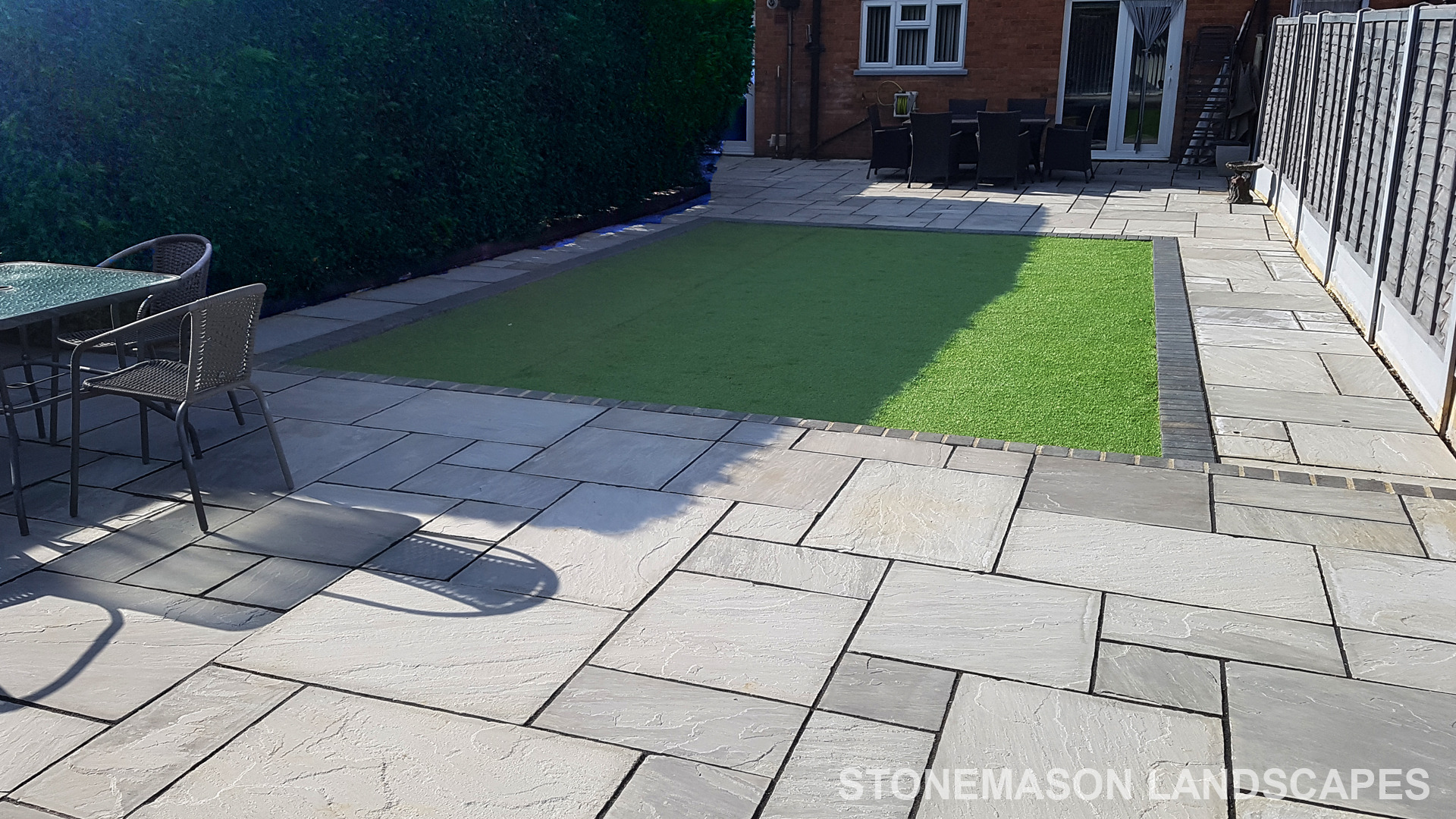 Fully paved grey sandstone patio with charcoal edging bocks and artificial grass lawn
