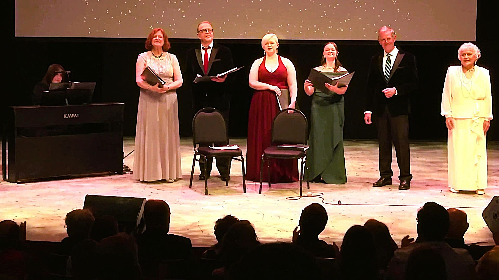 Singers in formal attire performing a vocal concert on stage with a piano accompanist.
