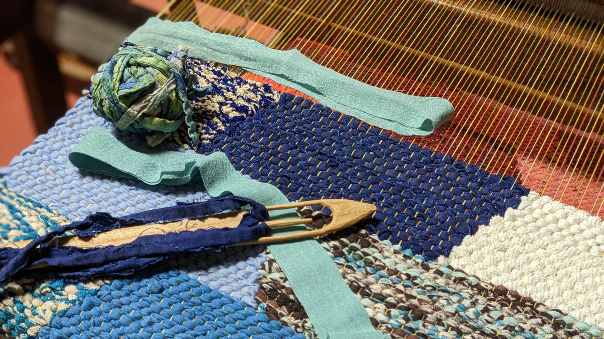 a weaving loom with a wooden shuttle, a ball of rag threads and a rag rug in shades of blue in progr