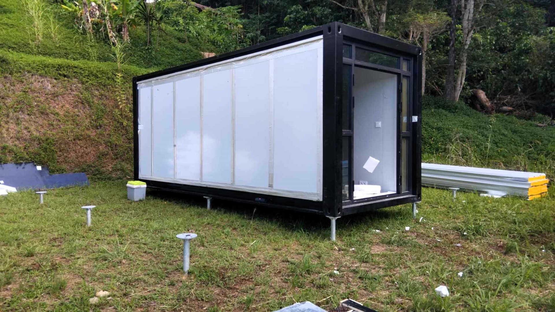 Maison conteneur noire préfabriquée sur pieux vissés dans un champ.