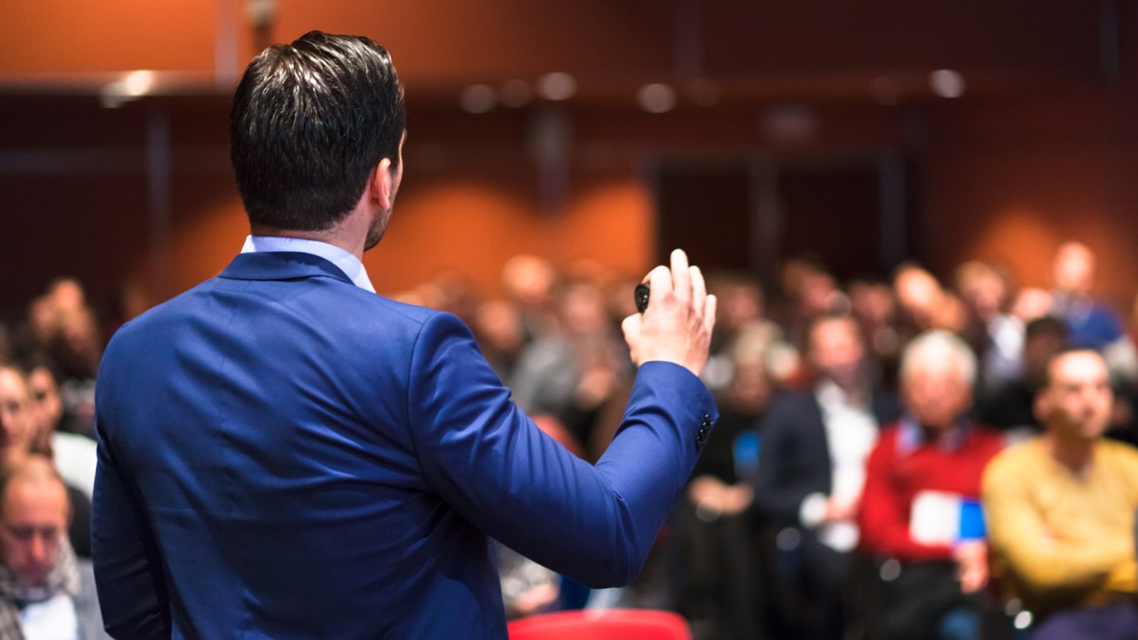 A keynote speaker in a suit presenting to a large audience at a conference.