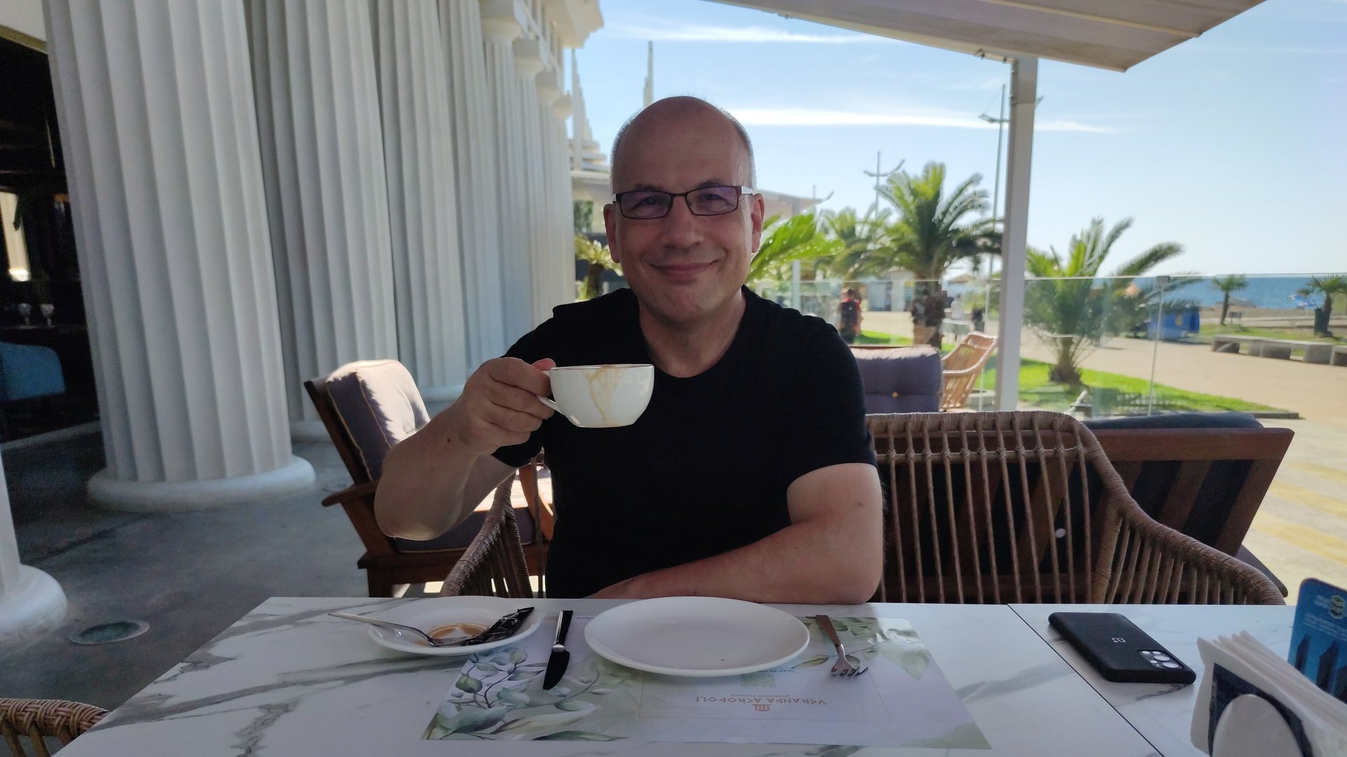 Smiling Heinz Roelfs in glasses enjoying coffee at an outdoor seaside terrace restaurant.